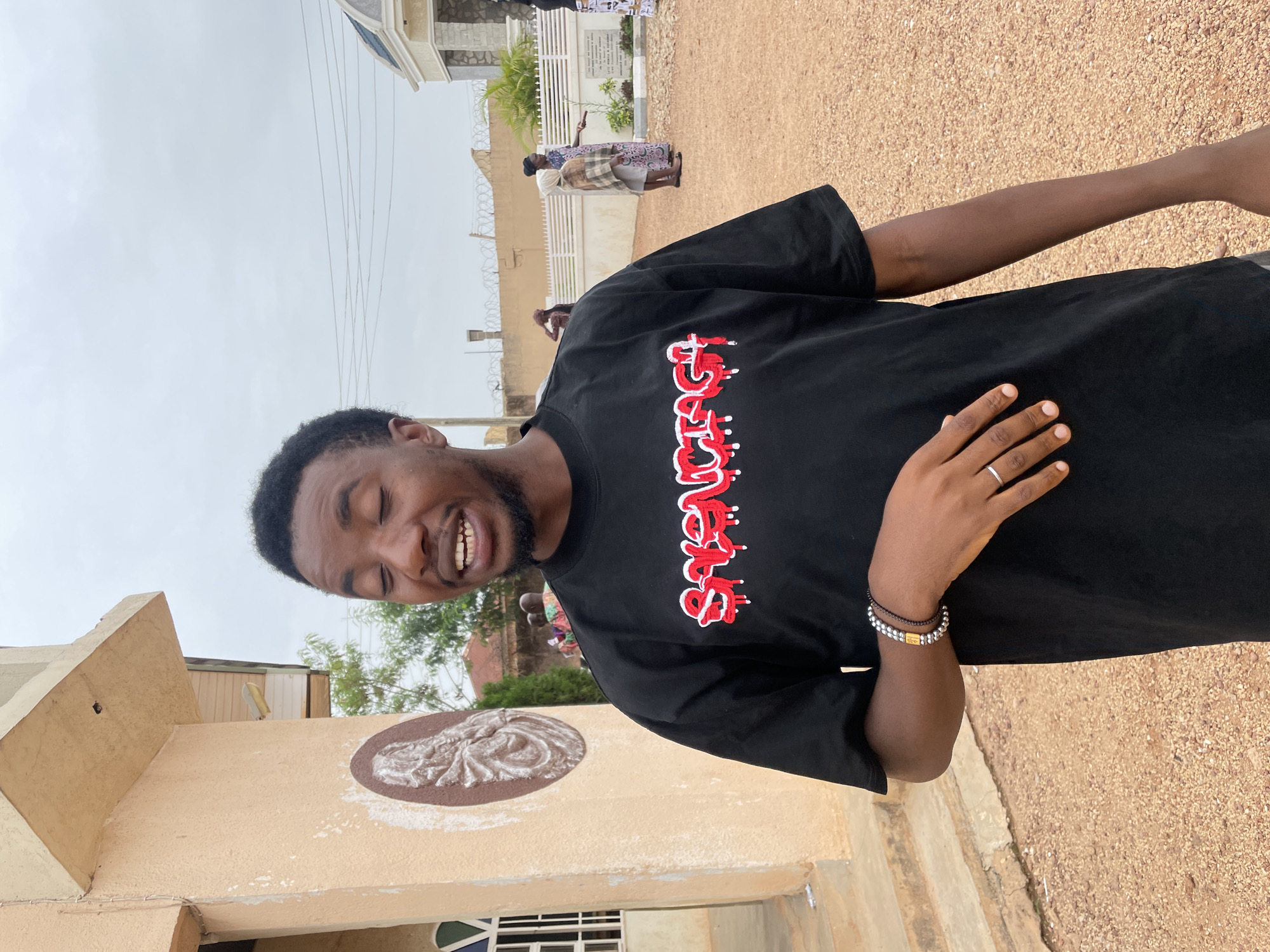 A black man is outside a sand-colored building in Jos, Nigeria. He is wearing a black t-shirt with white and red lettering, a silver ring, and a silver and black bracelet. He is smiling with his eyes closed. In the background, two women are standing on a stone walkway with a cemetery visible behind them. The ground is covered in tan gravel. The sky is overcast.