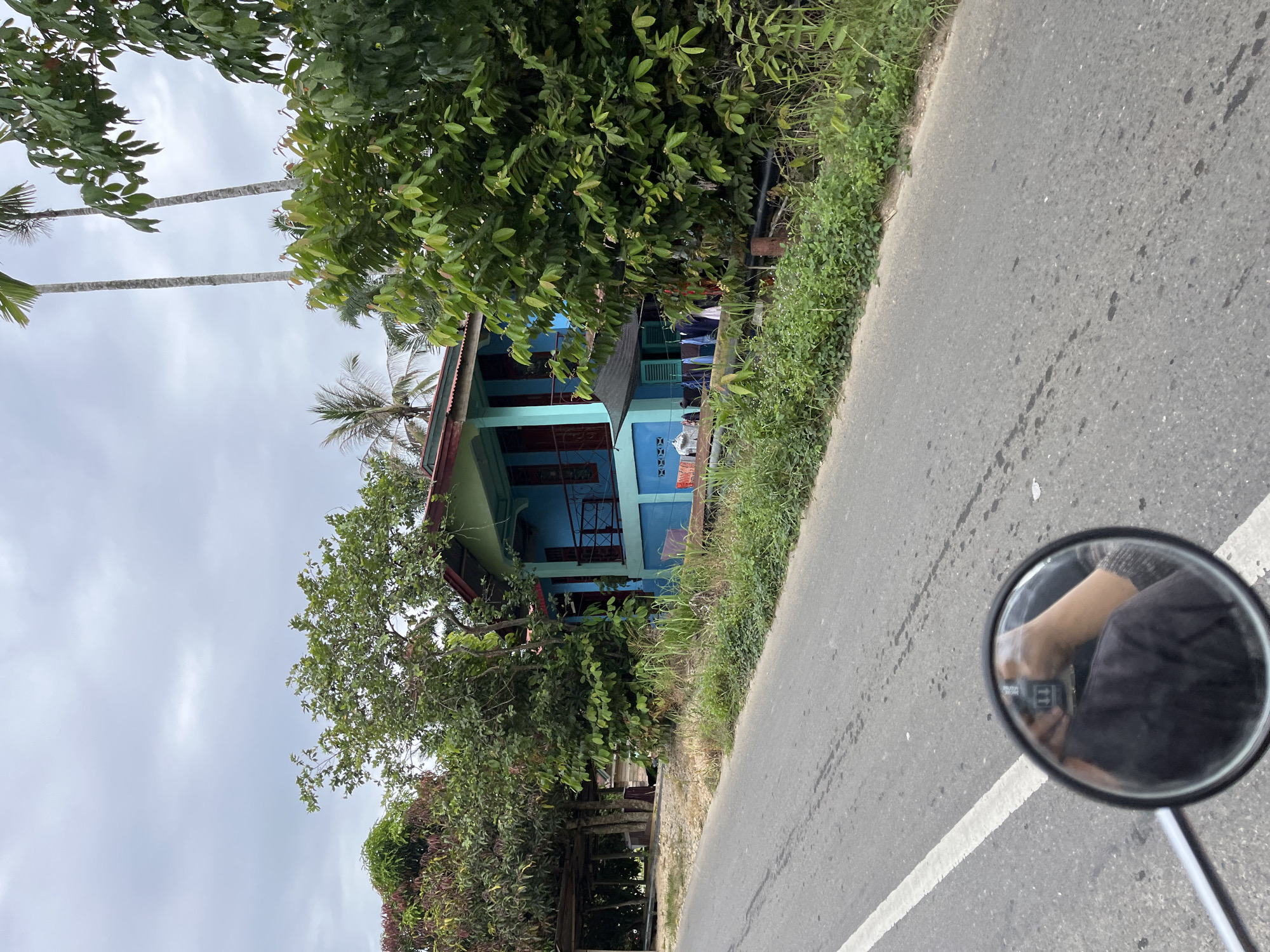 The image depicts a residential scene on a cloudy day in Sukaramai Dua, Indonesia. A two-story blue house with a green roof is situated amidst lush vegetation. The house has a balcony on the upper floor and laundry hanging outside on the lower level. A paved road runs in the foreground, with a white line marking the lane. A motorcycle's side mirror is visible in the bottom left corner, reflecting a person's arm and hand holding a mobile phone. The surrounding environment is characterized by dense trees, tall palm trees, and an overcast sky.