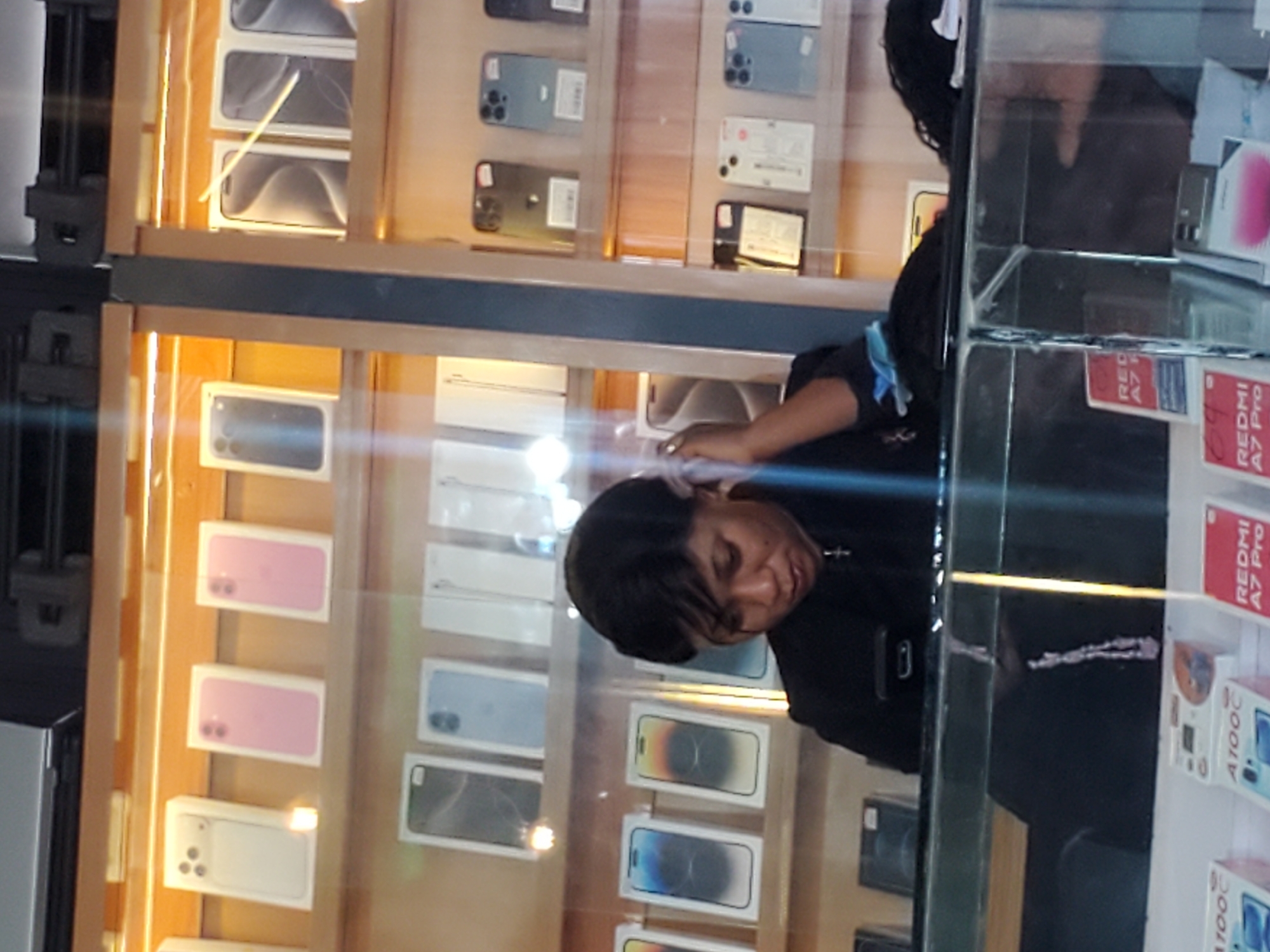 The image shows a close-up view of a display shelf in a store, likely a phone shop, with various mobile phones and their packaging arranged neatly. The lighting suggests it is daytime, possibly inside a well-lit establishment. The phones are displayed in boxes, some showing the phone itself through a clear window, while others are in opaque packaging with printed images. The boxes are stacked vertically and horizontally.
A person is partially visible in the foreground, leaning over the display. Their head and shoulder are visible, and they appear to be looking at the phones. Their hair is dark, and they are wearing a black top with a blue ribbon or tie. The person's face is not fully visible, but their posture suggests engagement with the products.
The setting appears to be a retail environment. The shelves are made of wood, and there are glass barriers or windows in front of the display. In the background, more shelves with phones are visible, creating a sense of abundance. Some of the packaging has text, including what appears to be "REDMI A7 Pro" and "A100C".
The overall impression is a busy retail environment where mobile phones are being showcased. The image is taken at an angle, with a slight blur, which could indicate movement or a quick snapshot. The focus is on the product display and the interaction of the partially visible individual with it.