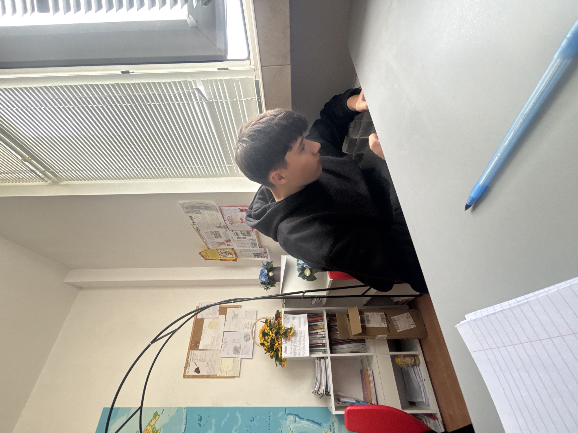 An indoor scene, likely a study room or office in Zlatograd, Bulgaria, captures a young person seated at a light grey desk during daytime. The individual, a male with short, dark hair, wears a black hoodie and is seen from the side, looking towards the right, seemingly focused on an activity at the desk. His right hand is visible, reaching for or interacting with something on the desk surface.
On the desk, a light blue ballpoint pen rests towards the right, and a sheet of lined paper is partially visible in the bottom right corner, suggesting work or study. To the left of the person, a window with white horizontal blinds allows bright daylight to flood the room, casting strong, distinct shadows.
The background features a white wall adorned with a cork board holding various pinned papers, some with illegible text and sketches. A large map, possibly a world map, is partially visible on the lower section of the wall. To the right, a white shelving unit holds an assortment of books, notebooks, and small cardboard boxes; one box has faintly visible numbers "3 15" on a label. Decorative elements include artificial bouquets of yellow sunflowers and blue hydrangeas. A lamp with a curved black arm is also present. The bright natural light indicates it is daytime, creating a clear and functional environment.