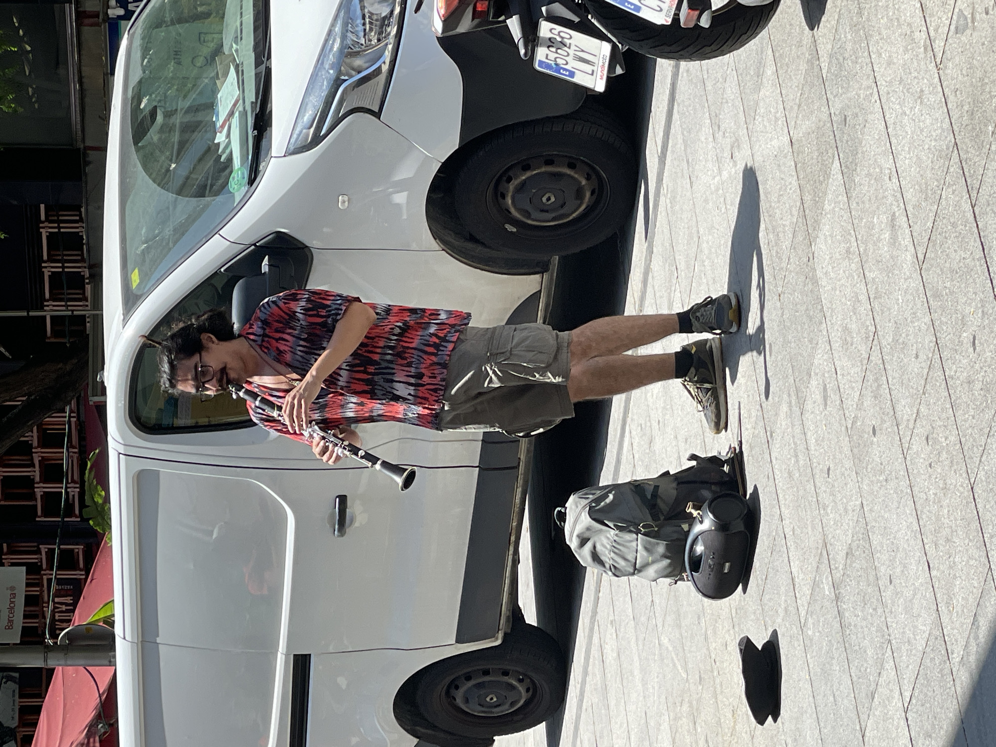 A man with dark hair, glasses, and a mustache is playing a clarinet on a sunny day in Barcelona, Spain. He is wearing a patterned short-sleeved shirt and khaki cargo shorts. He is standing next to a white van. In front of him on the ground are a gray backpack, a black portable speaker, and a dark-colored hat. A motorcycle is parked to the right of the van, with its license plate visible. The ground is paved with stone tiles.
