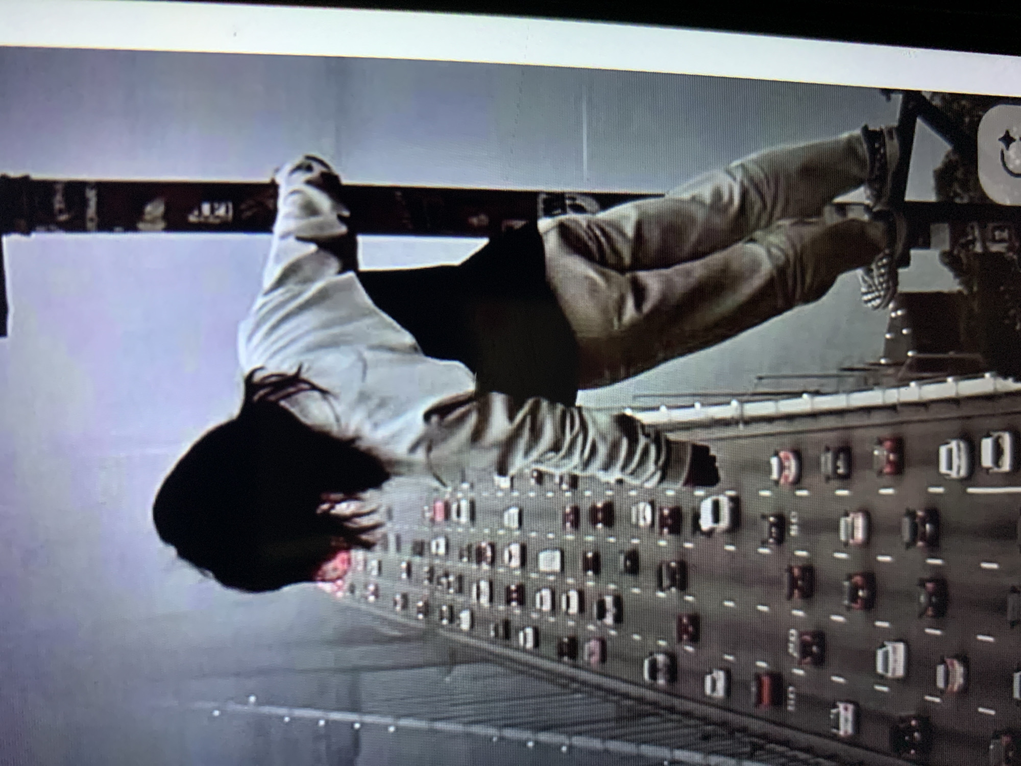 An image shows a person hanging from a pole on a bridge, with a heavy traffic jam below. The person is wearing a white shirt and beige pants, with black and white checkered shoes. The bridge appears to be a suspension bridge, with a long line of cars stretching out in front of it. The sky is overcast, and the scene is somewhat blurry, suggesting a low-resolution image or a significant distance from the camera. The specific location in the city of Lymanka, Ukraine is not discernible from the image itself.