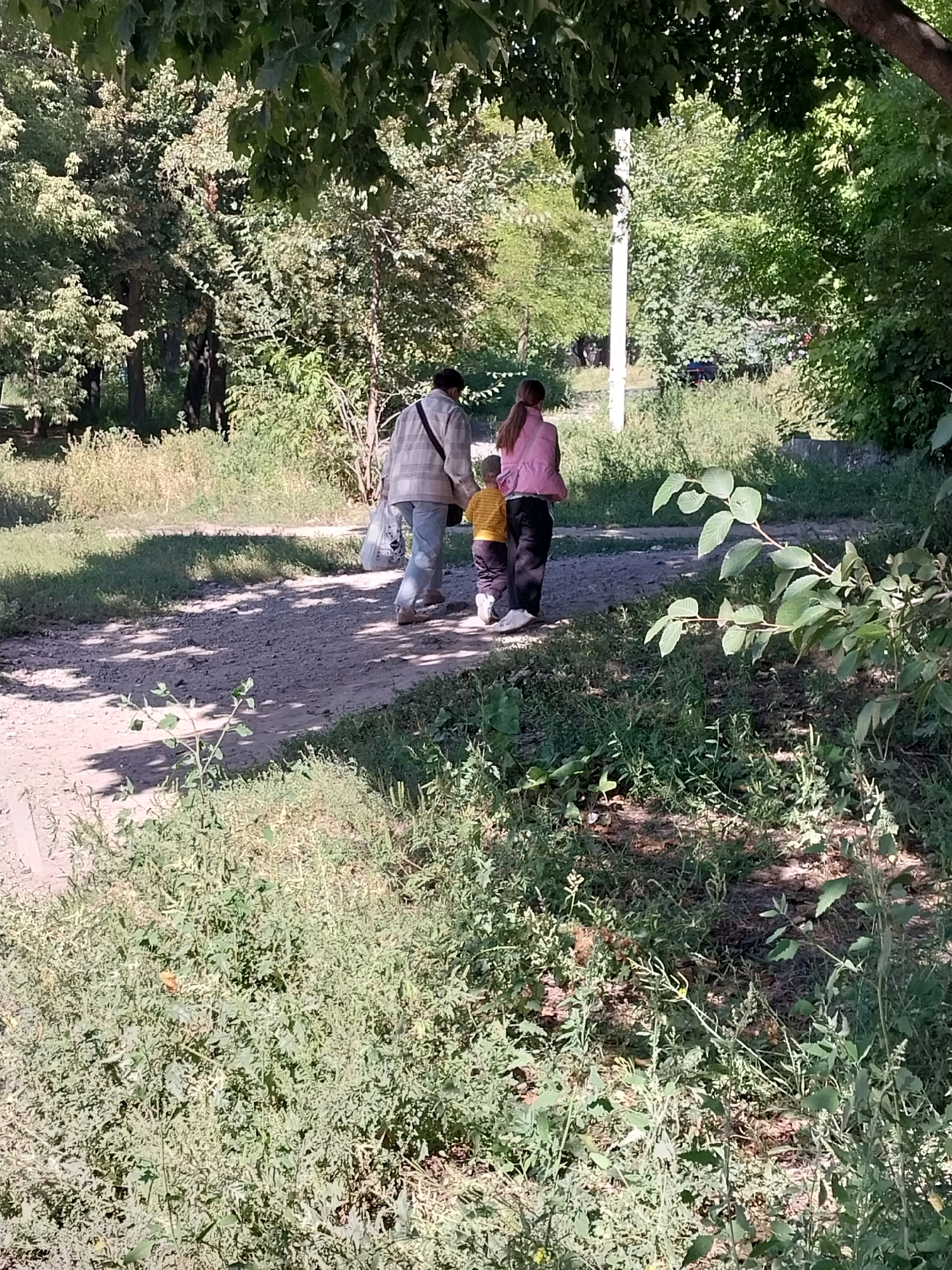 In a sun-dappled park area, three individuals are walking away from the camera on a dirt path. The person in the center, an adult, is wearing a plaid jacket and carrying a plastic bag. To their right is a child in a yellow shirt and dark pants. To the left of the adult is another child wearing a pink jacket and dark pants. The path is bordered by overgrown grass and trees, with a white pole visible in the background. The scene is consistent with a public park within the city of Dnipro, Ukraine.