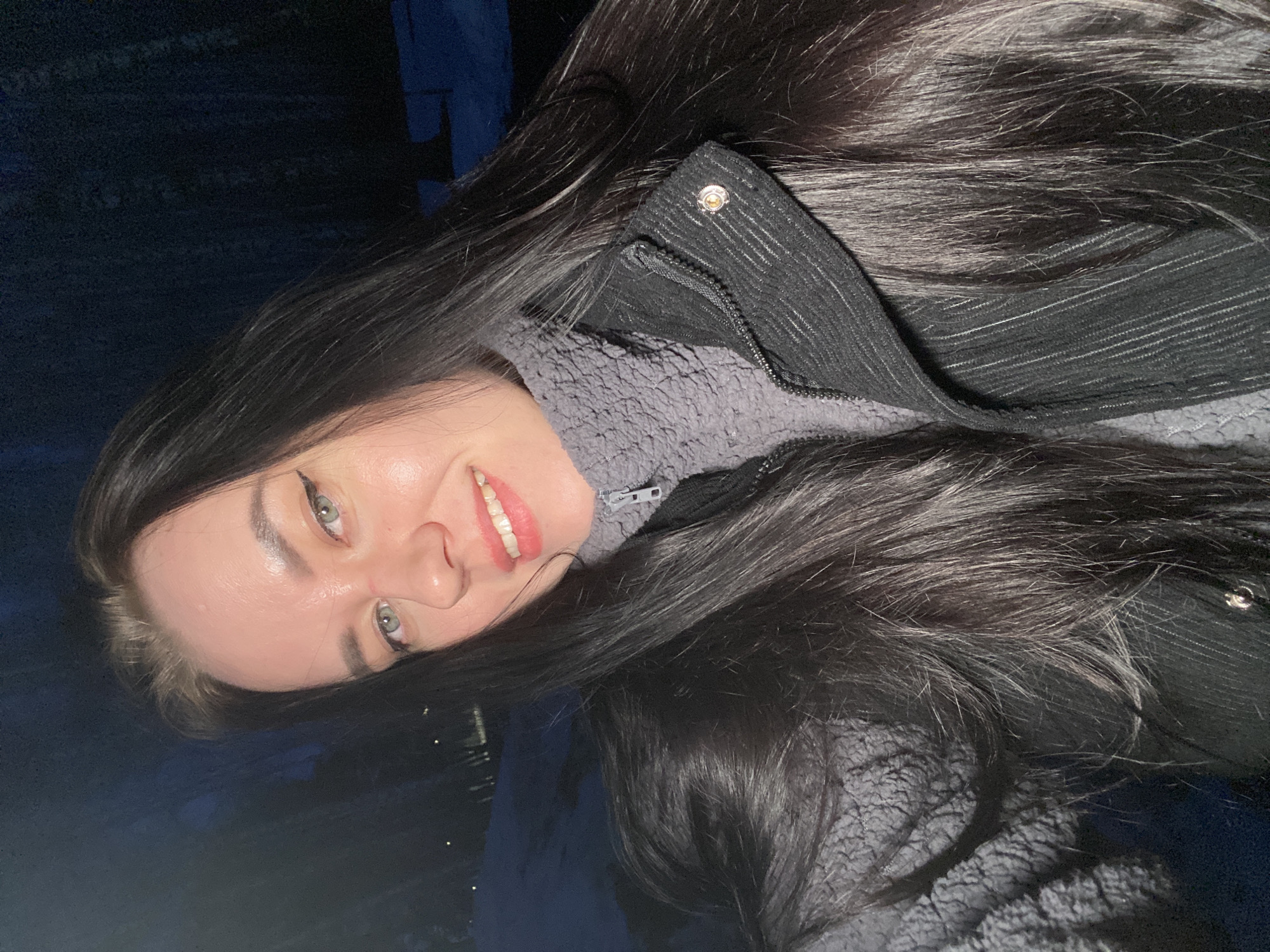 A young woman with long, dark hair and light eyes smiles at the camera. She wears a gray fleece jacket layered under a black jacket with a silver button detail near the collar. The background appears dark and indistinct, possibly suggesting an outdoor setting at night. It is not possible to verify location in the city of Chaikovsky, Russia.