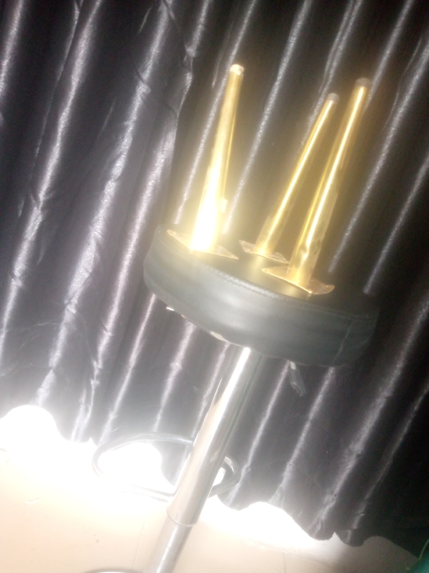 The image displays an indoor scene, location in the city of Unknown, Unknown. A black, upholstered bar stool with a chrome-plated base and footrest is centrally positioned. Four slender, highly reflective golden-colored metallic poles, approximately of equal height, are affixed vertically to the top surface of the stool's seat. Each golden pole has a flat, square-shaped base where it attaches to the stool. The background consists of dark, vertically pleated curtains with a visible sheen, suggesting a satin or similar reflective fabric. The bottom left foreground reveals a portion of a light-colored floor, significantly overexposed by a bright light source emanating from that area, which also causes strong glare and reflections on the metallic components of the stool and the golden poles. No individuals are present, and no actions or interactions are depicted.