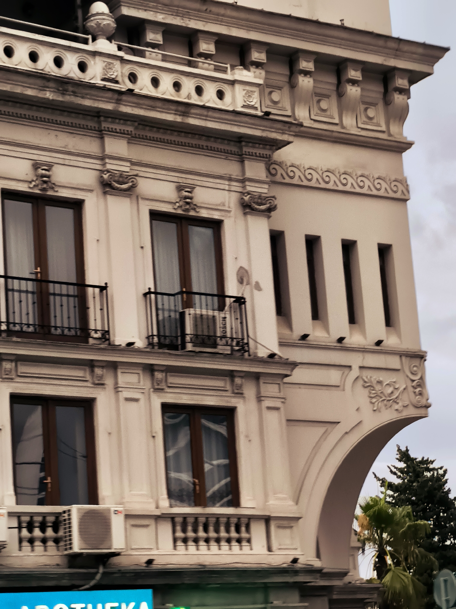 The image shows a close-up, exterior view of the upper floors of a light-colored, ornate building in Batumi, Georgia. The architecture features classical elements such as pilasters, decorative moldings, balconies with wrought-iron railings, and arched openings. Several windows with dark wooden frames and curtains are visible. On one of the balconies, an air conditioning unit is mounted. The sky is overcast. In the lower part of the frame, a bright blue sign with white lettering that reads "APOTHEKA" is partially visible. Green foliage, including palm trees and a coniferous tree, can be seen peeking out from behind the building on the right side.