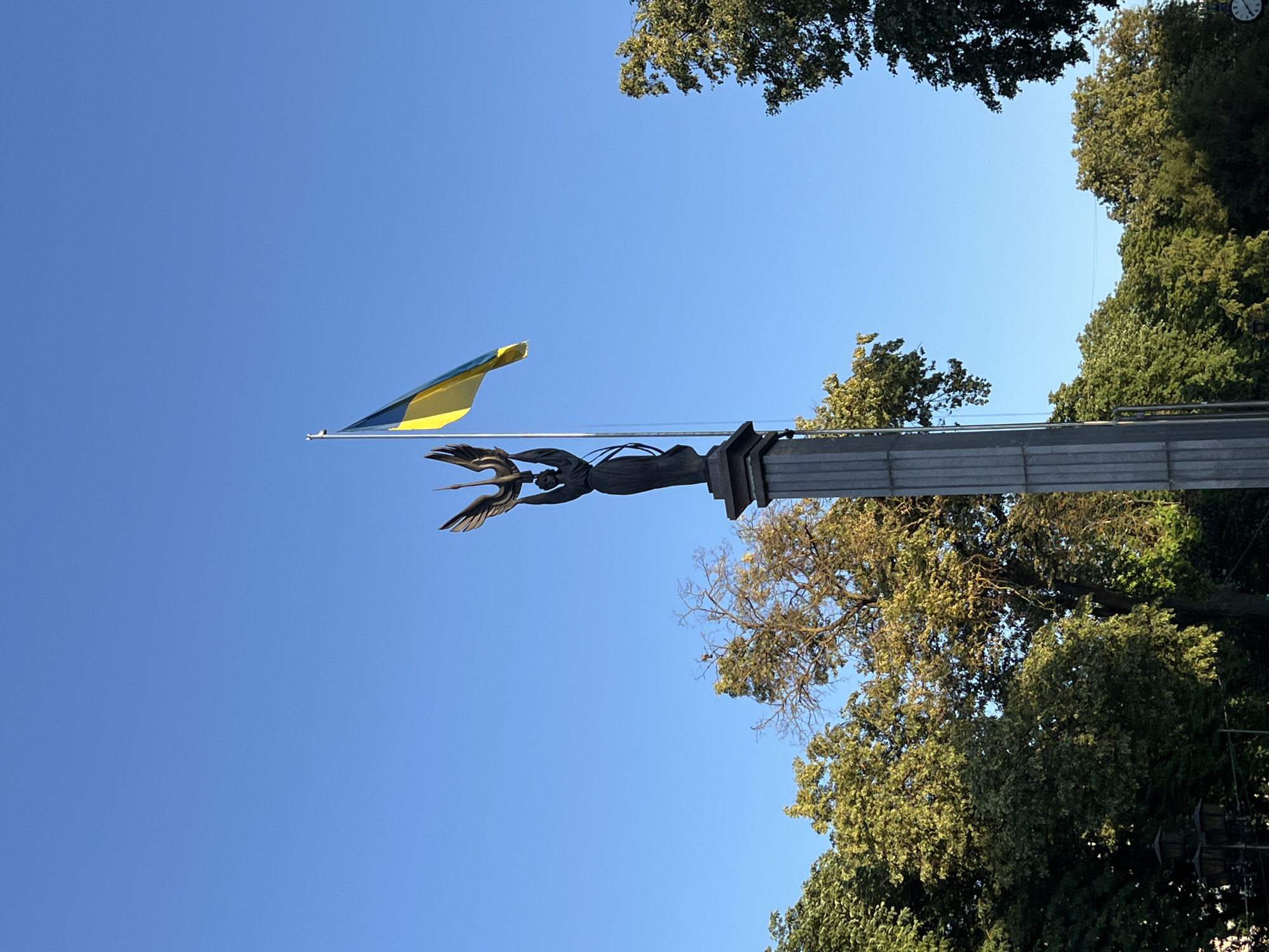 The image depicts a Ukrainian flag, blue and yellow, fluttering from a flagpole. The flagpole is adorned with a dark-colored sculpture of a winged figure, possibly an angel or a mythological creature, with its arms outstretched. The sculpture is mounted on a stone monument.
In the background, lush green trees form a canopy against a clear blue sky, suggesting an outdoor setting on a bright, sunny day. The perspective is from below, looking upwards towards the flag and the trees. The composition is framed by the branches and leaves of the trees, creating a sense of enclosure.
The scene appears peaceful and patriotic, with the national flag prominently displayed. There are no people visible in the frame. The time of day is likely daytime, given the bright sunlight illuminating the scene and casting shadows on the trees. The location is identified as Ternopil, Ukraine, adding a specific geographical context to the image. There is no visible text in the image.