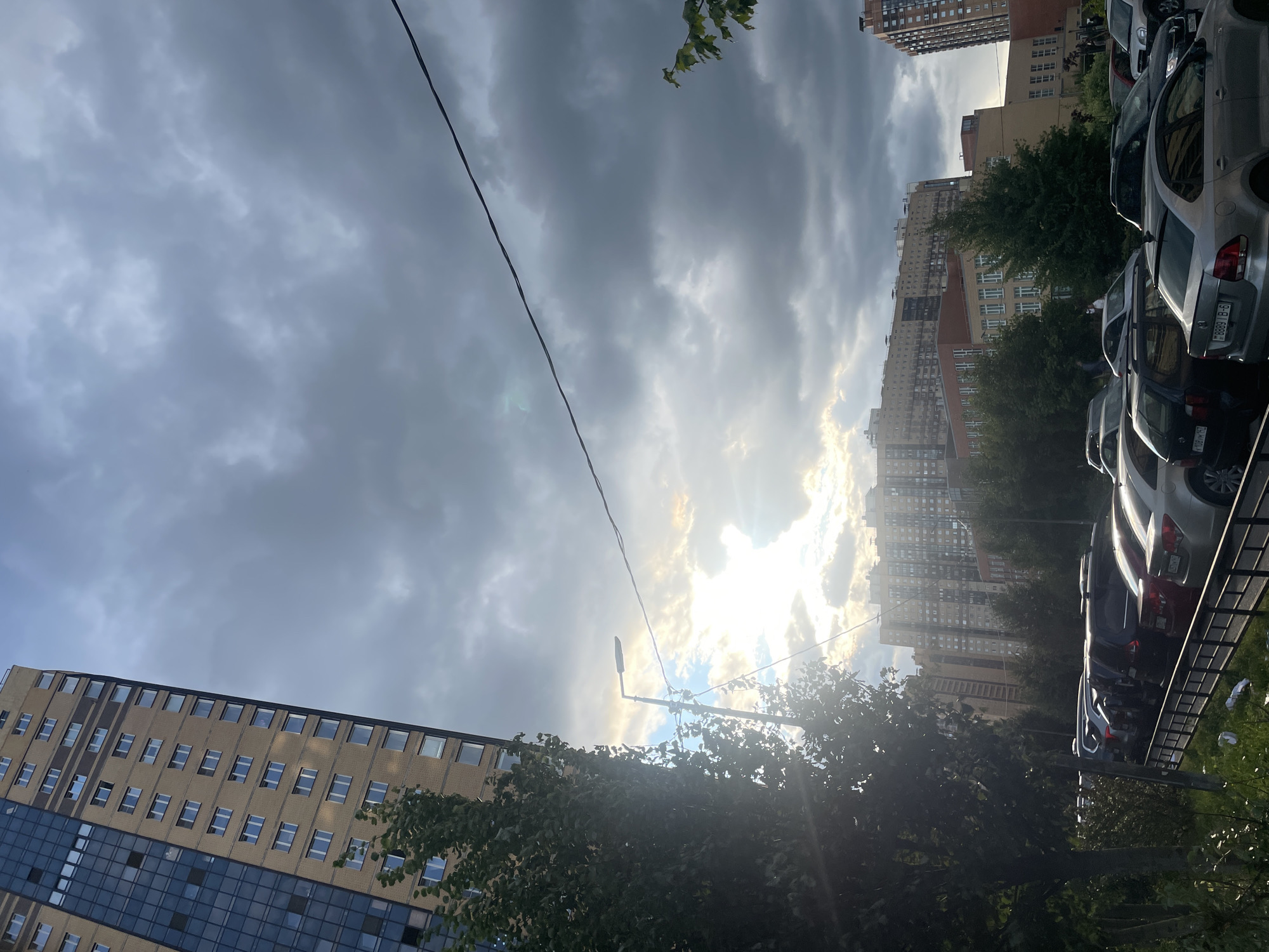In Ладожское Снт, Russia, the image captures an outdoor urban scene under a cloudy sky. A tall, multi-story residential building with a beige facade and dark-tinted windows dominates the left side of the frame. A large tree with lush green foliage is positioned in front of the building, partially obscuring it and casting shadows. In the mid-ground, a row of parked cars is visible, including several sedans and SUVs. Behind the cars, a long, multi-story building with a light-colored facade and numerous windows can be seen, with trees interspersed between it and the foreground. Further in the distance, other residential buildings are visible, some with dark, tinted windows and others with lighter facades. The sky is filled with dramatic, dark grey clouds, with patches of sunlight breaking through in the upper right and center, creating a somewhat diffused and atmospheric lighting effect. A street light stands on a pole near the tree in the mid-left of the image, and a power line runs diagonally across the sky from the upper right to the mid-left.