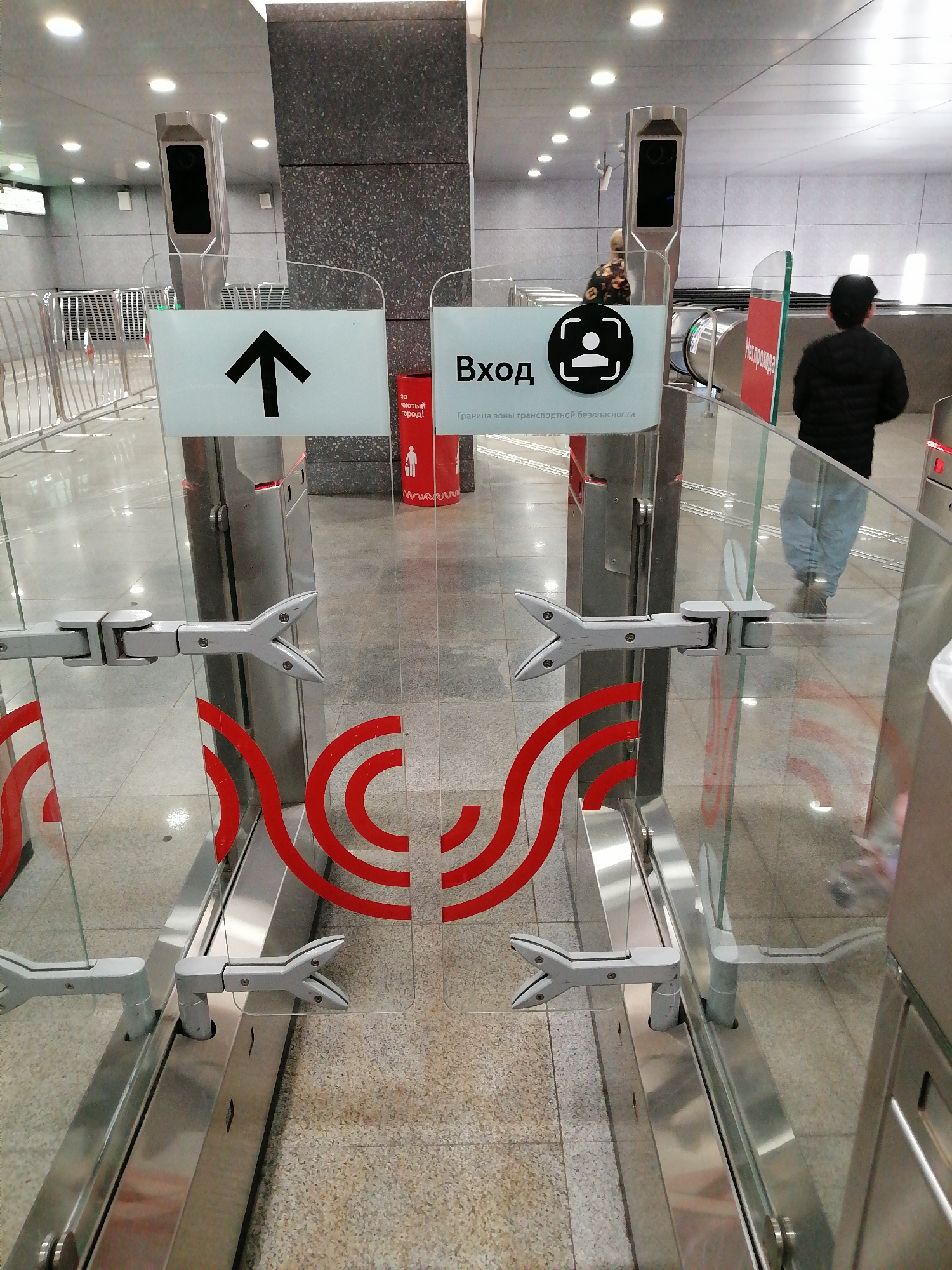 The image shows a turnstile at a metro station in Moscow, Russia. The turnstile has glass barriers with a red curved design. Signs indicating entry and direction are posted above the gates. A person wearing a black jacket and blue jeans is walking through the turnstile. In the background, escalators and other people can be seen. The flooring is a light-colored tile.