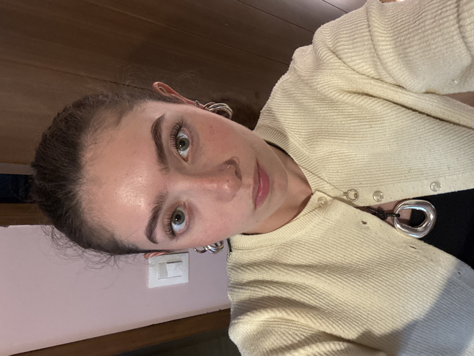 A young woman with light skin, brown hair pulled back, and green eyes is shown in a close-up, angled shot. She wears silver hoop earrings and a nose ring. A cream-colored, buttoned cardigan covers a black top, and a silver pendant necklace is visible. The background consists of a pink wall with a white light switch and a section of a brown wooden surface.