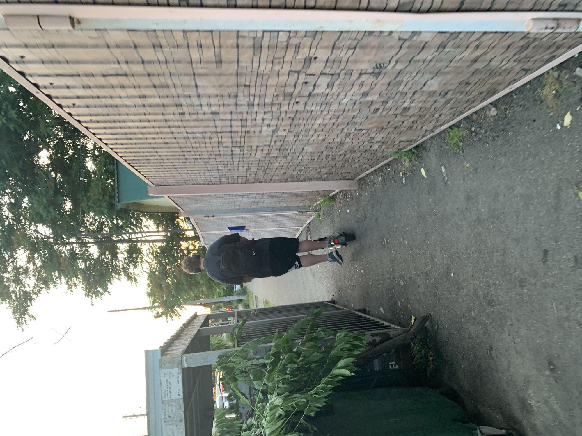 In the city of Unknown, Unknown, a young person wearing headphones and a backpack rides a scooter down a narrow paved path. The path is bordered on the left by a green dumpster and a low, covered structure with visible graffiti. On the right, a tall, wooden fence with a stone-like pattern stretches into the distance. Trees and foliage are visible in the background, and the sky is bright, suggesting it is either early morning or late afternoon.