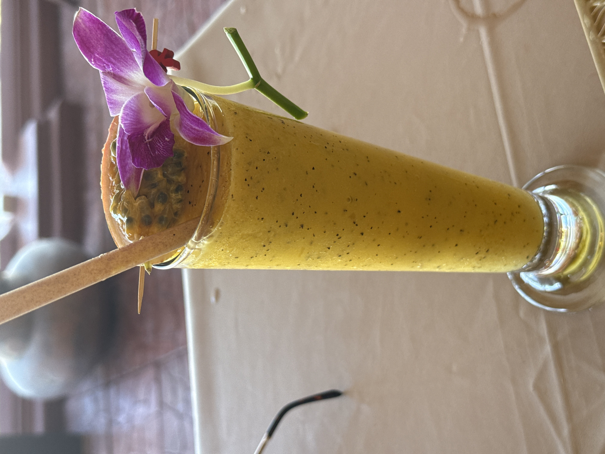 A tall, clear glass filled with a vibrant yellow, speckled smoothie is presented on a light beige, textured surface. The smoothie is garnished with a slice of passion fruit on top, secured by a wooden skewer, and a delicate purple orchid with a green stem. A thin, light brown bamboo straw is inserted into the smoothie. In the background, out of focus, is a round, light gray object and a wooden surface. To the left of the glass, a dark object with a curved arm is partially visible, possibly glasses. The overall scene appears to be set in an outdoor dining area in Baan Lamai, Thailand, given the tropical flower and lighting.
