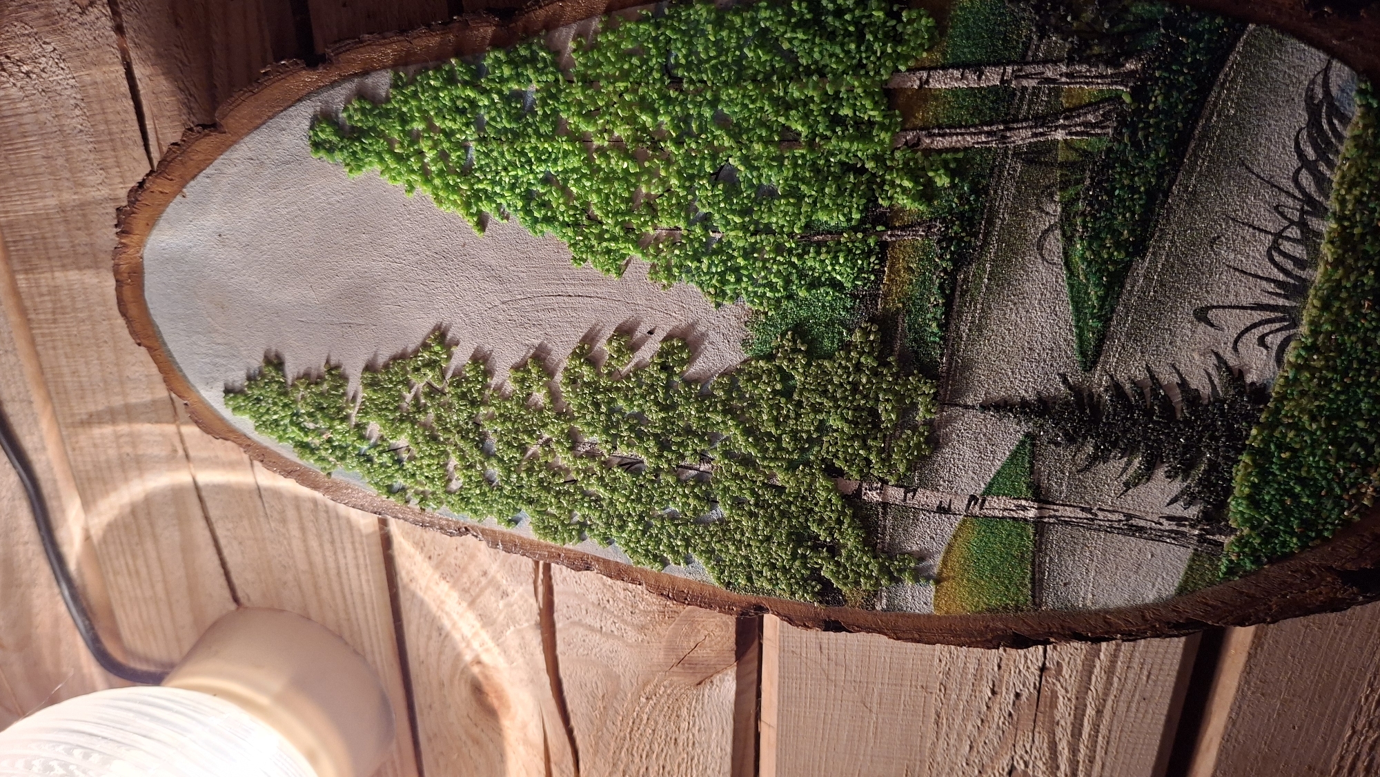 The image depicts a decorative wooden plaque with a landscape scene. The plaque is oval, with its bark edge visible. The scene features trees rendered with what appears to be textured green material, possibly small beads or moss. Between the "trees" is a gray background depicting mountains. The plaque is positioned against a background of wooden planks, and a light fixture is partially visible in the lower left corner. The scene appears to be indoors. The location is Большой Букор, Russia, as specified in the prompt.
