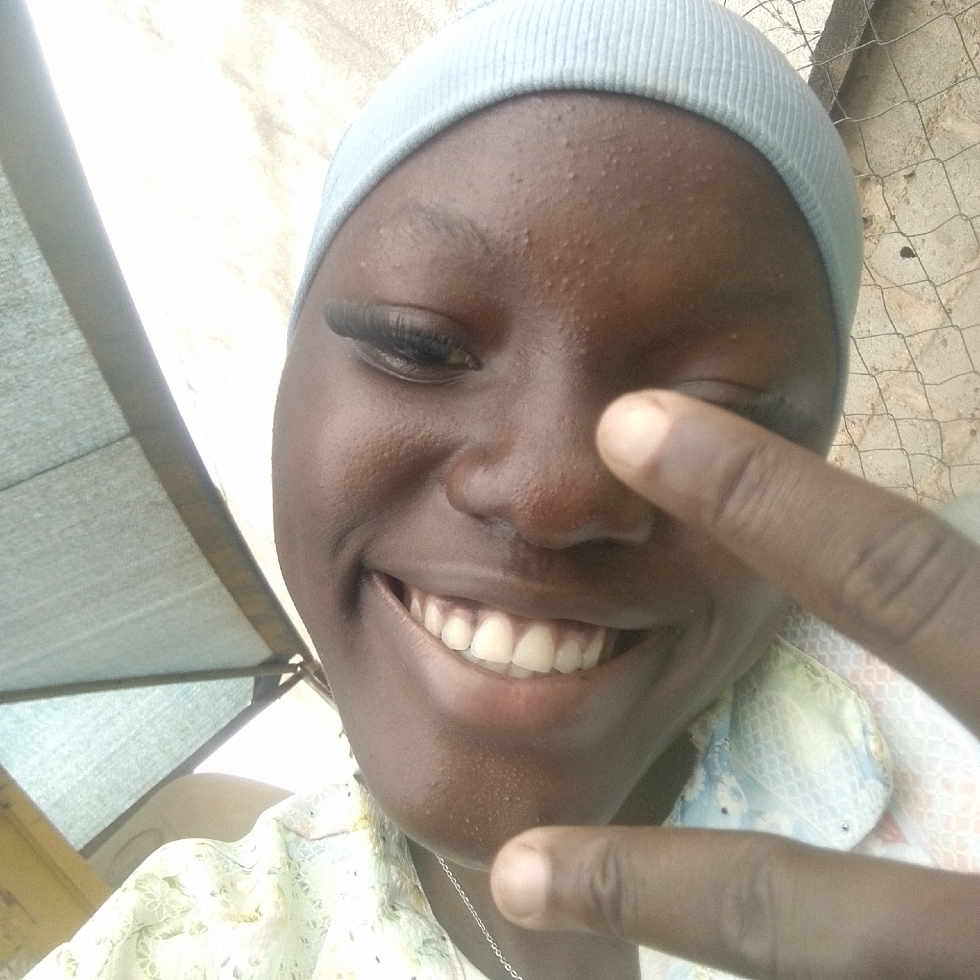 A close-up, low-angle selfie taken outdoors features a young person wearing a light blue beanie. The individual has dark skin and a visible smile, revealing white teeth. Their right eye is partially covered by their index finger, which is extended towards the camera. The background is blurred, showing a textured, light-colored wall and a wire fence. To the left, a light-colored tarp or awning is visible, casting a shadow. The person is wearing a light-colored shirt with a floral pattern. The overall lighting suggests it is daytime.
