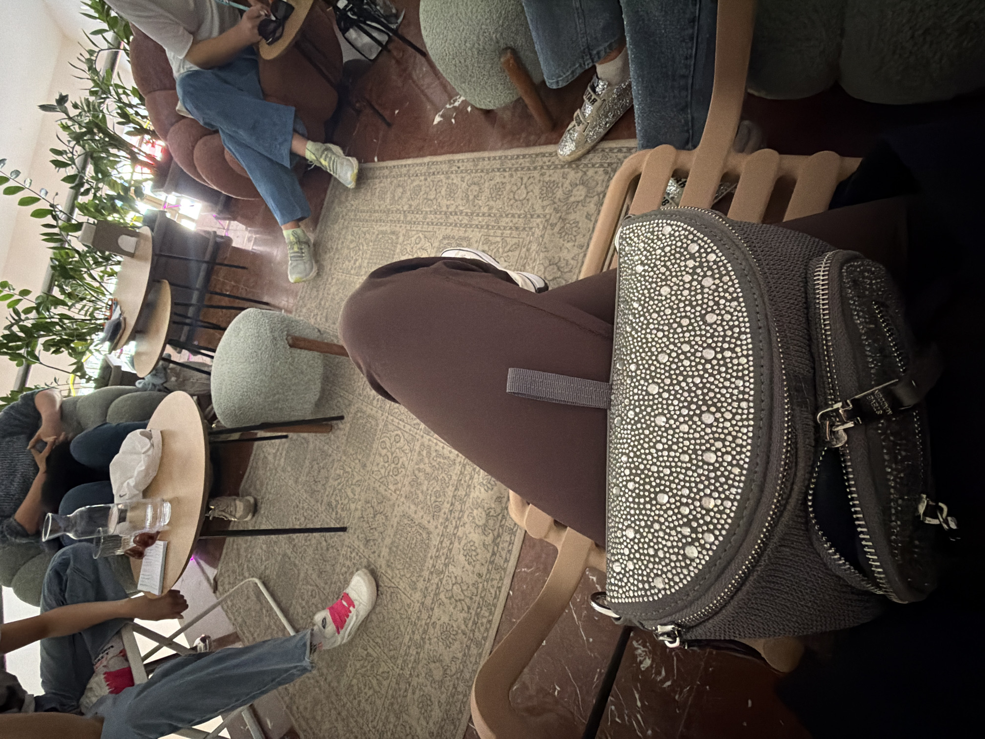The image depicts a casual indoor gathering in a bar or cafe in Bar, Montenegro. Several individuals are seated and interacting in a relaxed setting.
In the immediate foreground, a dark grey bag covered with numerous silver studs or rhinestones is prominently displayed, with the brand name "STEVE MADDEN" visible on its zipper pull. A person next to the bag is wearing brown pants and a white sneaker. Further into the scene, other individuals are visible, mostly from the waist down. One person wears blue jeans and white sneakers with bright pink accents, while another wears blue jeans and light-colored sneakers with yellow details, seated on a rounded brown armchair. Another person is seen wearing blue jeans and distinct sparkly silver sneakers. Some individuals are holding phones, indicating casual engagement or communication.
The setting features a mix of furniture, including round wooden tables, upholstered green stools, and comfortable armchairs. A light-colored rug with a detailed pattern covers much of the floor. Green leafy plants are placed around the room, enhancing the cafe aesthetic. On one table, a clear glass bottle of water, a glass, and a small bowl containing red items (possibly berries or cherries) are visible, along with a white cap. The atmosphere appears calm and social, suggesting a leisurely meeting among friends or acquaintances. The lighting is ambient and typical of an indoor space, without strong indications of specific time of day other than a general brightness.