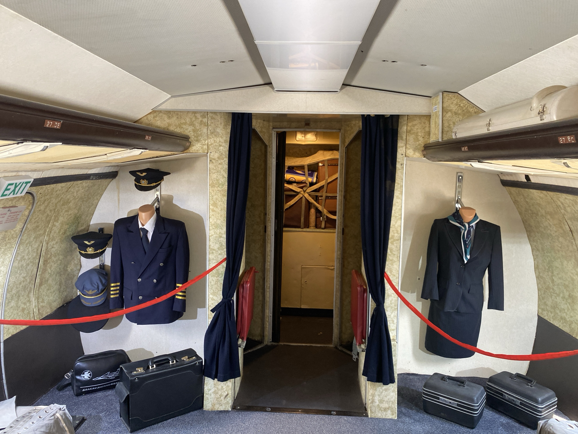 The image depicts the interior of a display simulating an aircraft cabin, possibly part of an exhibition at the Museum of Aviation in Burgas, Bulgaria. On the left, a pilot's uniform is displayed on a mannequin, complete with a dark blue jacket with gold stripes on the sleeves, a white shirt, a dark tie, and two caps (one black with gold insignia, and another blue with gold trim). Two black suitcases are also on the floor to the left of the mannequin. To the right, a flight attendant's uniform is displayed, consisting of a dark suit jacket, skirt, white shirt, and a patterned scarf. Two dark grey train cases are on the floor in front of this mannequin. A narrow doorway with dark blue curtains on either side leads into another section of the aircraft, showing a glimpse of the interior with some cargo secured. Overhead compartments are visible, with one on the left labeled "27.DE". A red rope barrier is positioned in front of the uniforms and luggage, indicating a viewing area. The overall environment suggests a preserved or recreated section of an airplane, set up for public viewing.