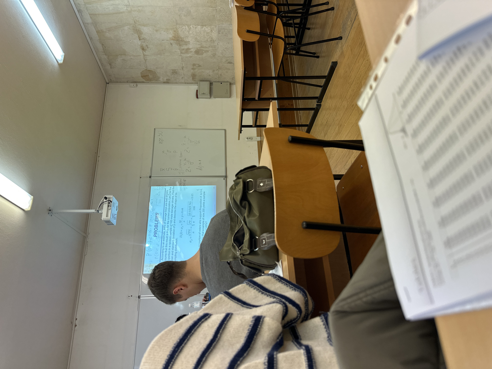 A student is attending a statistics lecture in a classroom. The student is looking at a projector screen displaying statistical problems. A whiteboard behind the screen has mathematical equations written on it. The location is most likely in Varna, Bulgaria, at a university, based on the type of lecture. The classroom has wooden desks and chairs. Other students are also present. The image focuses on the student in the foreground with a backpack. The scene is brightly lit with fluorescent lights.