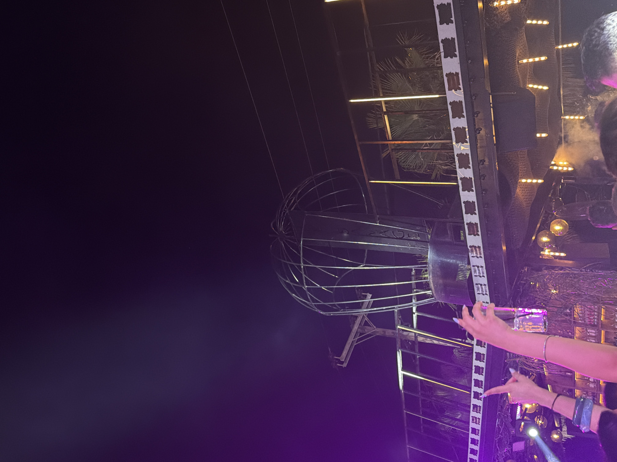 The image depicts an outdoor nighttime scene, likely a club or entertainment venue, illuminated by purple and yellow lights. In the foreground, two arms are raised, one holding a glass and the other pointing. The background features a prominent metallic structure resembling a large, open cage or dome, positioned above a balcony with palm trees visible behind it. The overall atmosphere suggests a lively social gathering, possibly in the city of Kassandra Municipal Unit, Greece, known for its nightlife.
