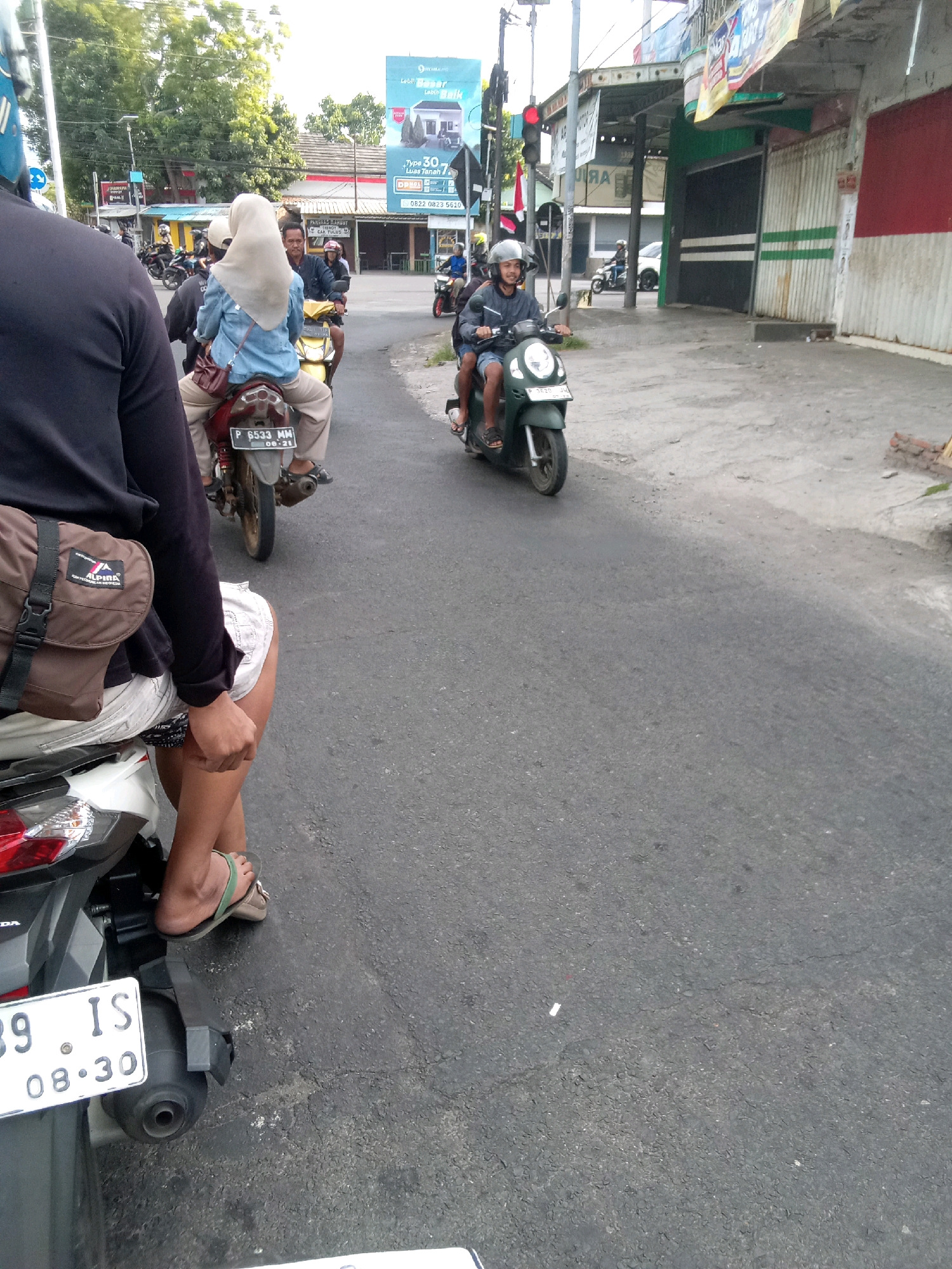 The image captures a street scene in Jember, Indonesia, with several motorcycles and riders on an asphalt road. In the foreground, a person is riding on the back of a motorcycle, wearing a dark long-sleeved shirt and white shorts, with a brown bag slung over their shoulder. Their feet are visible, wearing flip-flops. Ahead of them, another motorcycle carries a rider wearing a blue jacket and a light-colored hijab.
Further down the road, a person in a grey jacket and helmet is riding a green scooter. Traffic lights are visible in the background, with a red light illuminated. Buildings line the street, some with advertisements and shops. A large blue billboard with text and images is also visible, promoting housing with the text "Lagi Pasang type 30 Luas Tanah." Other motorcycles are seen in the distance, suggesting moderate traffic. The overall environment appears to be an urban or semi-urban area, likely during daylight hours, given the lighting conditions.