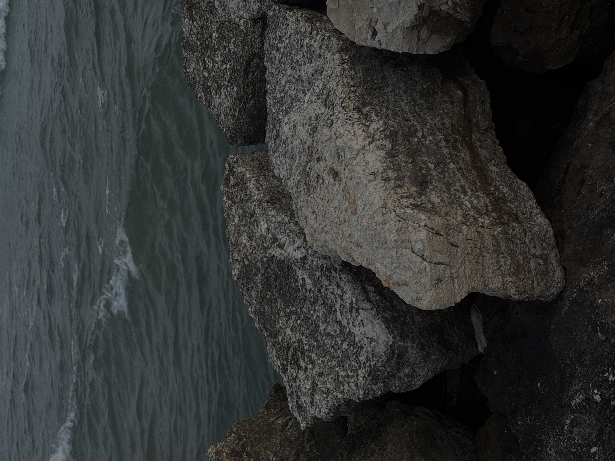 The image shows a view from above, looking down at a rocky shoreline and the sea. The left side of the frame is dominated by gray, choppy water. The right side is composed of large, rough-hewn rocks, stacked together. The rocks vary in shade from light gray to almost black, creating dark shadows between them.
There are no people visible in the scene. The location is likely in Tel Aviv, Israel, as coastal areas are present and similar rock structures can be found along the city's coastline.