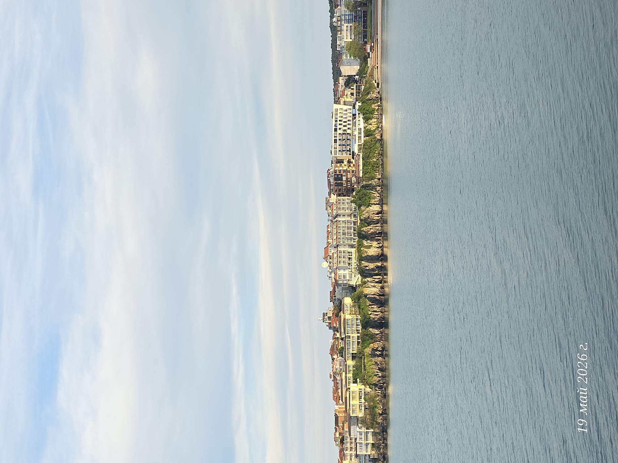 The image shows a coastal scene, likely taken in Sozopol, Bulgaria, on May 19, 2026. The sky is mostly blue with thin, elongated clouds. On the right is the sea, its surface showing subtle ripples and a uniform, light blue color. Buildings line the coastline, perched on rocky terrain with patches of greenery. The architecture appears to be a mix of residential and tourist accommodations. The light suggests it is daytime, with the sun positioned in a way that it illuminates the building facades. The overall atmosphere seems calm and peaceful.