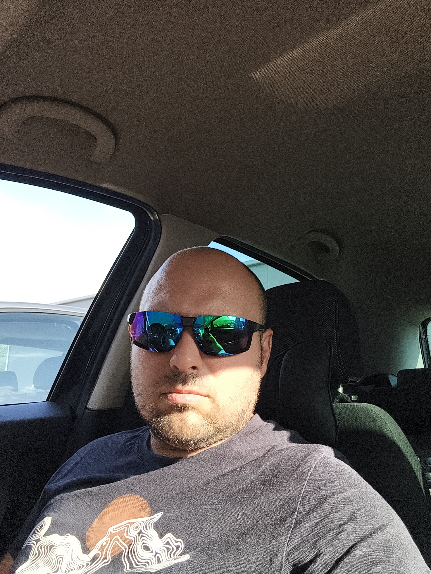 A man is sitting in the driver's seat of a car. He is bald and wearing sunglasses. He is also wearing a dark t-shirt with a graphic on it. The car is parked in an outdoor setting, possibly a parking lot, with the sky visible through the windows. The interior of the car is dark, with some light coming in from outside. There is a handle above the driver's side door and the passenger seat is visible behind the driver. The man is looking directly at the camera. The lighting suggests it is daytime.