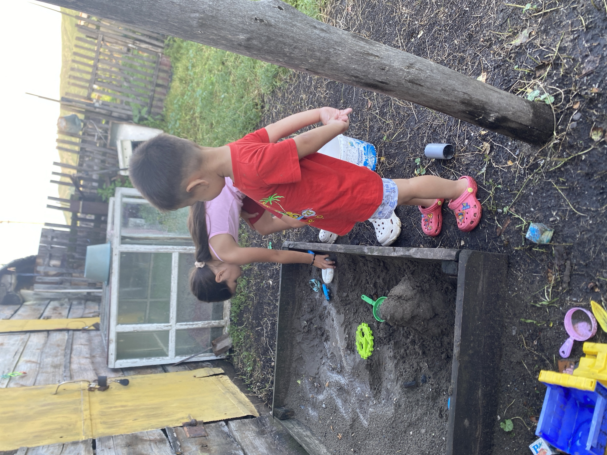 In Maloarslangulovo, Russia, two children, a girl and a boy, are engaged in play within a wooden sandbox. The girl, wearing a pink top and red shorts, is bent over the sand, seemingly interacting with toys. The boy, in a red t-shirt and patterned shorts, stands beside her, also facing the sandbox. Various sand toys, including a green gear-shaped mold and a green shovel, are scattered in the sand. In the background, a rustic wooden structure with a yellow door and a white-framed greenhouse are visible. A weathered wooden fence encloses the area, and distant hills are discernible under a bright sky. The ground around the sandbox is covered in dark soil and sparse vegetation.