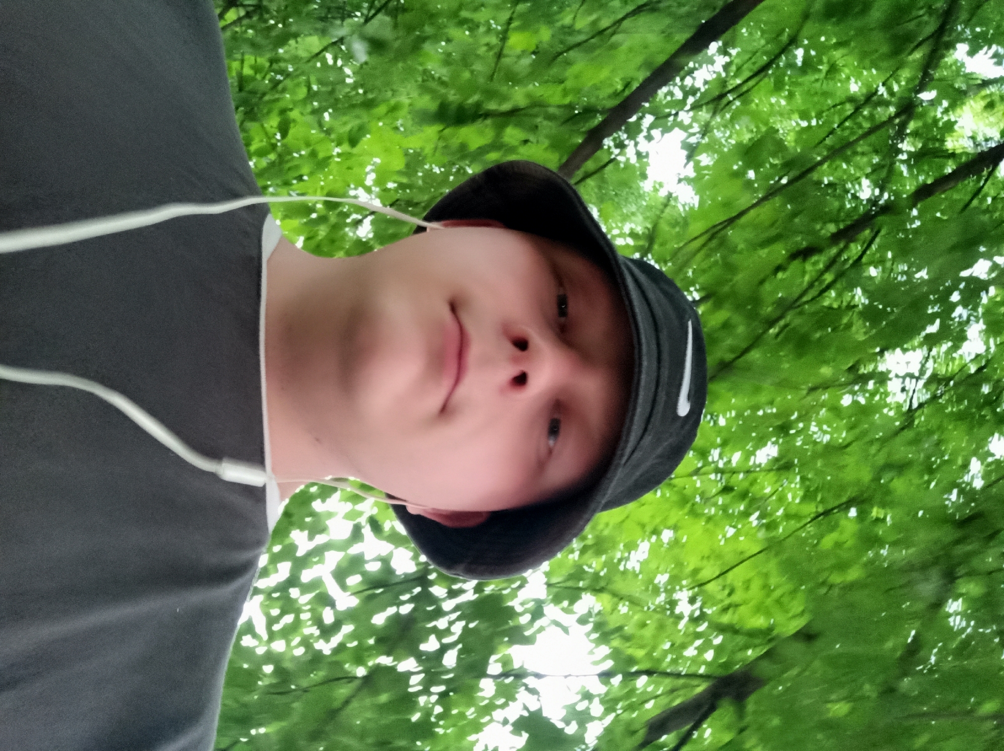 A person is shown from the chest up, wearing a dark bucket hat and a dark grey t-shirt. White earbuds are visible, with wires extending down. The person is looking upwards, with a neutral expression. The background is filled with lush green leaves and tree branches, suggesting an outdoor environment, possibly a park or a wooded area. The lighting is bright, with sunlight filtering through the leaves, creating a dappled effect. The image appears to be a selfie, taken from a low angle.