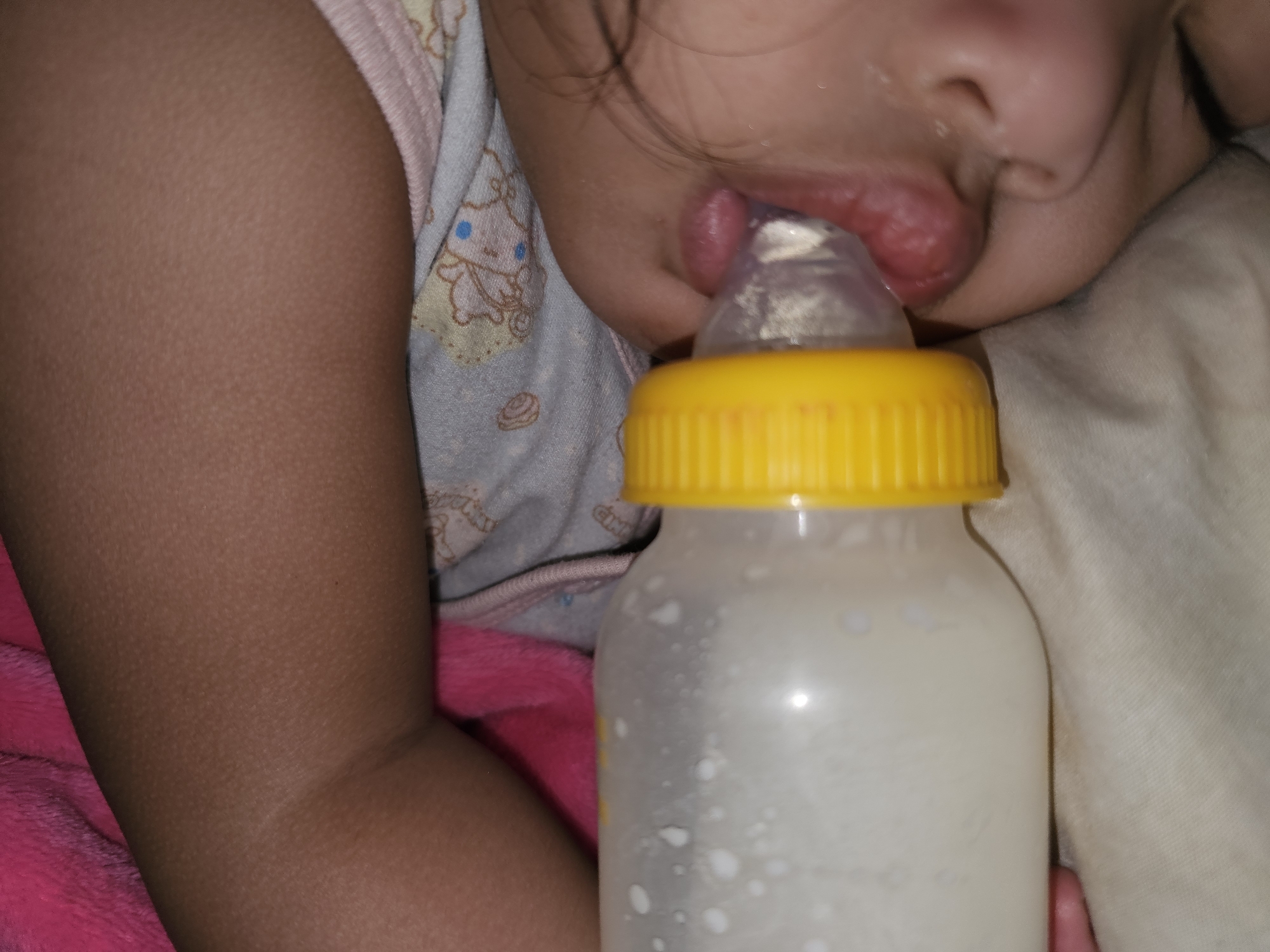 A close-up, slightly angled shot shows a baby's face and mouth feeding from a clear baby bottle with a yellow cap. The bottle is filled with a white liquid, possibly milk, with small bubbles visible inside. The baby's lips are slightly parted around the bottle's nipple. The baby is wearing a light-colored onesie with a teddy bear pattern. The background includes a soft pink blanket and a light brown fabric. The image appears to be taken indoors, in the city of Sumberwudi, Indonesia.