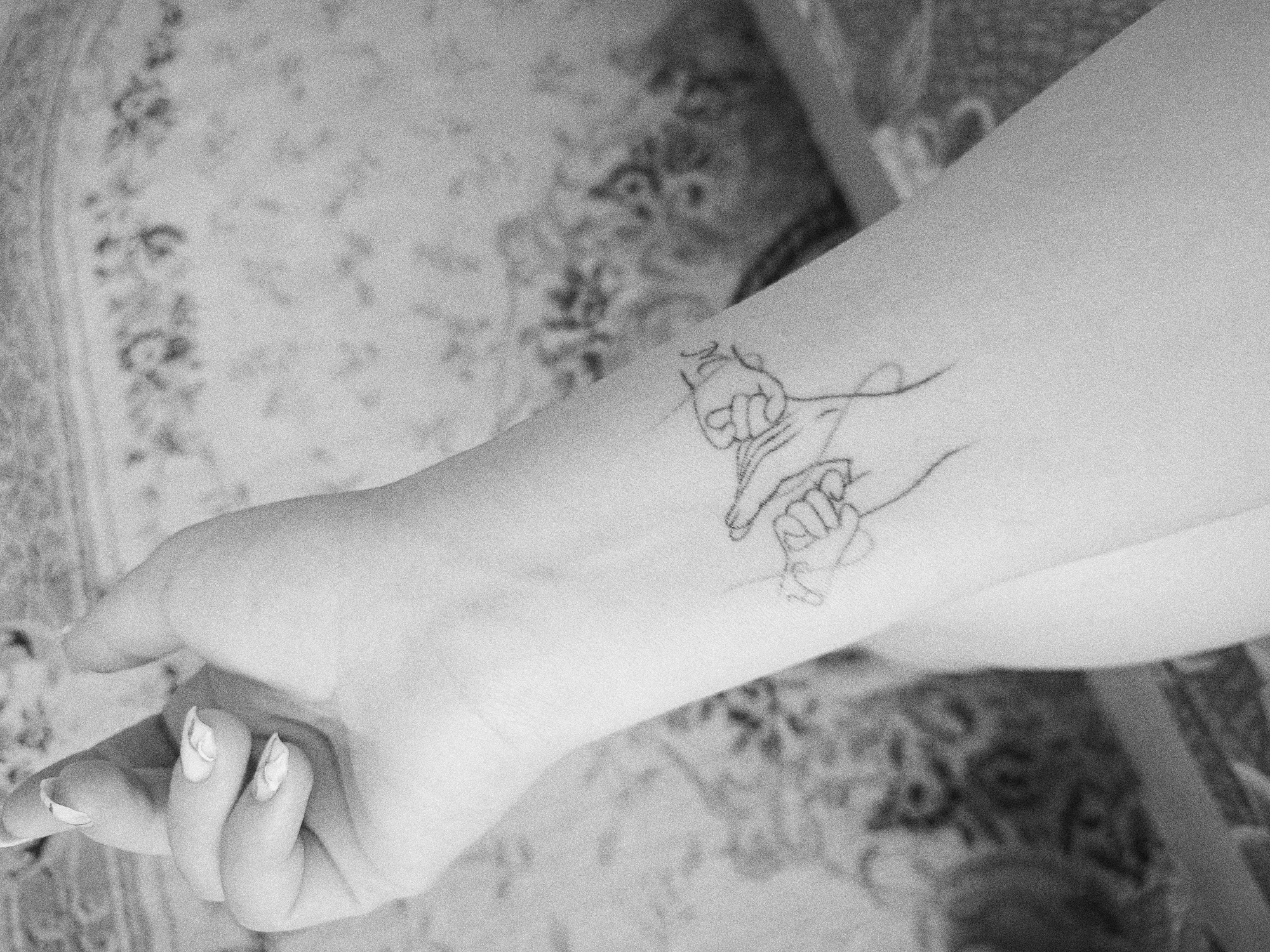 A black and white photograph shows a person's arm and hand with a simple line drawing tattoo on their forearm. The tattoo depicts two hands, possibly interlocked or connected. The fingernails on the person's hand are manicured with a light color and a subtle design. The arm rests on a surface with a patterned fabric. The location is not identifiable as Tobolsk, Russia based on the image alone.