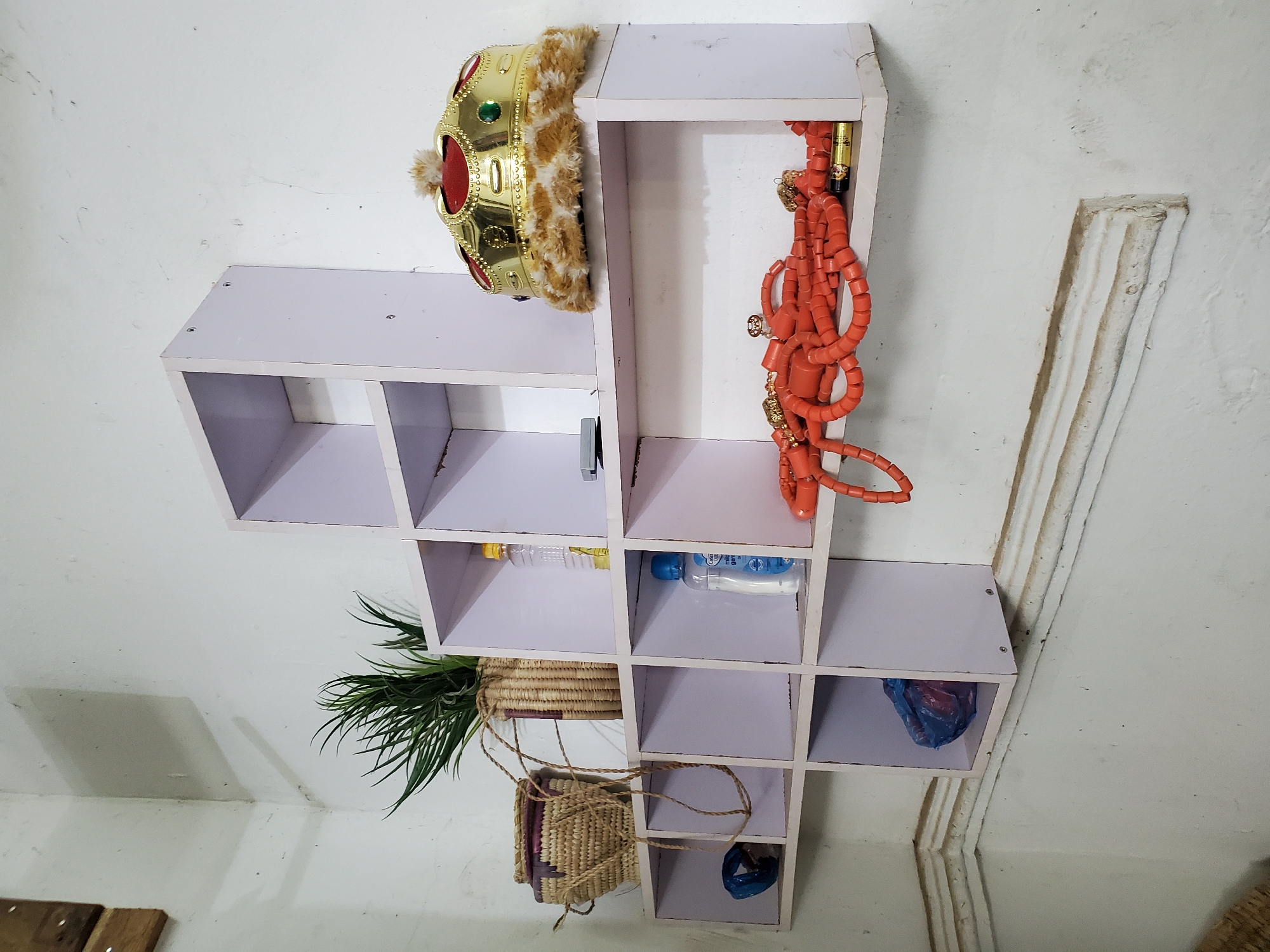 The image shows a white, cube-shaped shelf system mounted on a white wall. The shelf contains a gold-colored crown with fur trim, an orange beaded necklace, two bottles of liquid, a small potted plant, and two woven baskets. One of the baskets appears to contain a small blue object. The location is Mararaba, Nigeria.