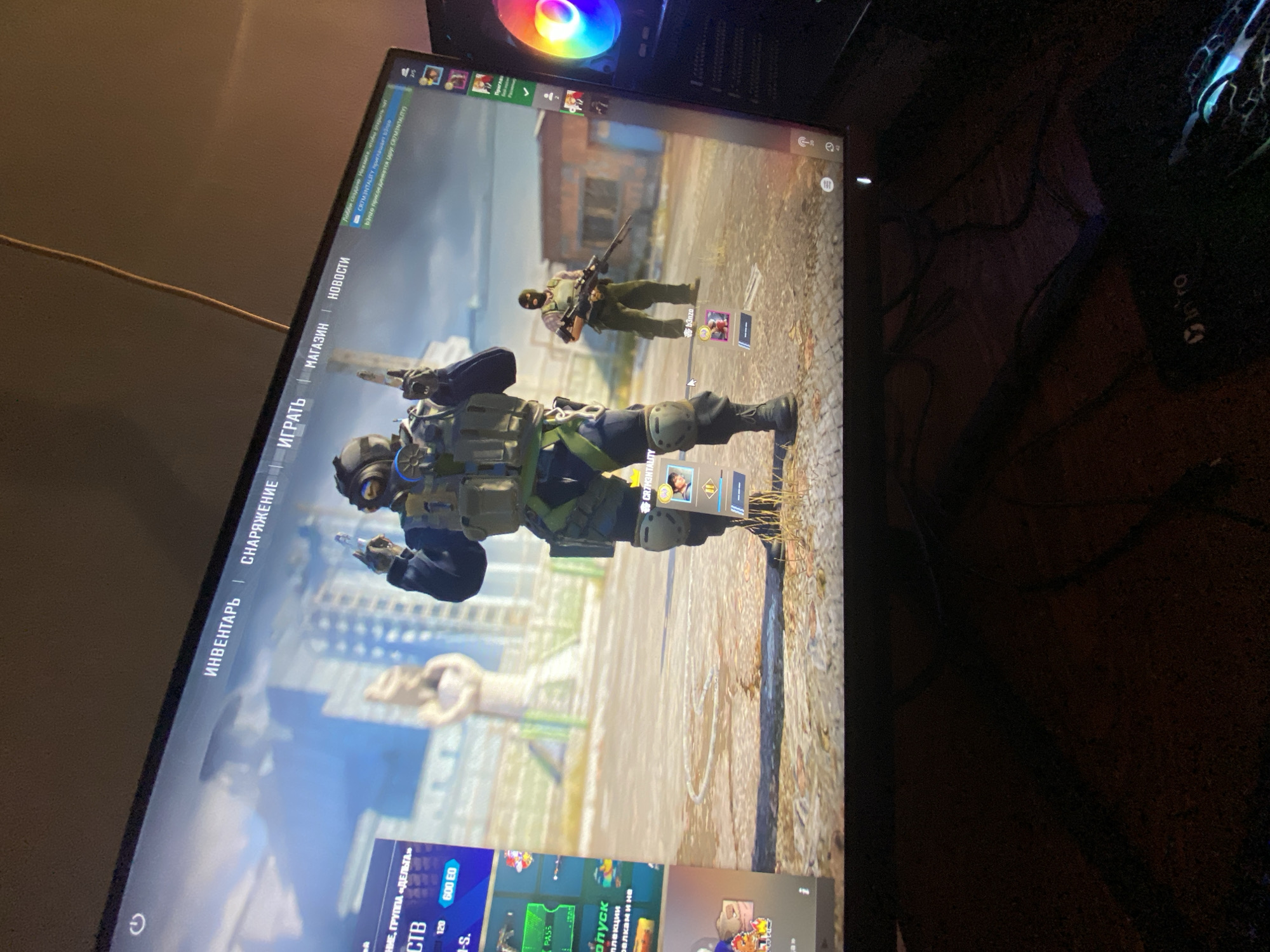 The image captures a computer monitor displaying a video game, likely in a room in Yerevan, Armenia. The monitor is tilted and reflects some ambient light. On the screen, a game interface is visible, featuring a main character in tactical military gear, holding a pistol, and a secondary character in a balaclava holding a rifle further in the background. The game's setting appears to be an urban environment with dilapidated buildings and a dirt or concrete ground.
The game's user interface is predominantly in Russian, with visible tabs and notifications. Key text elements include "ИНВЕНТАРЬ" (Inventory), "СНАРЯЖЕНИЕ" (Equipment), "ИГРАТЬ" (Play), "МАГАЗИН" (Shop), and "НОВОСТИ" (News), suggesting a game lobby or menu screen. Notifications like "Лобби создано" (Lobby created), "CR7M3NTALITY приглашает" (CR7M3NTALITY invites), and a mention of "b3nzo" indicate a multiplayer or social aspect of the game. Other game-related text includes "ГРУППА «ДЕЛЬТА»" (Group "Delta"), "CTB", "600 EO", and "PASS".
To the right of the monitor, part of a computer tower with a vibrant, rainbow-colored RGB fan is visible. Further to the right and slightly out of focus, a gaming mouse with the text "intro" and an Xbox logo can be seen. Various cables are tangled on the surface around the computer setup, suggesting a typical gaming station. The overall scene depicts an active gaming session, with the game characters and UI being the central focus.