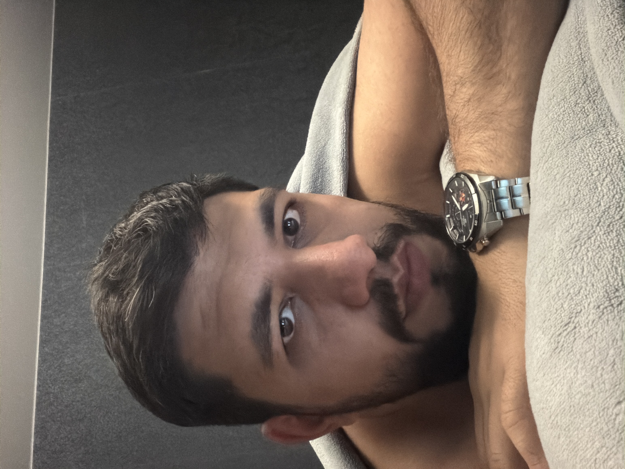 A man is lying down on a gray towel with his arm raised, displaying a silver wristwatch. The background consists of a dark gray wall on one side and a light gray vertical panel on the other. The man has short, dark hair with graying temples, brown eyes, a beard and mustache. There is no information available in the image to determine its location in Krasnoyarsk, Russia.