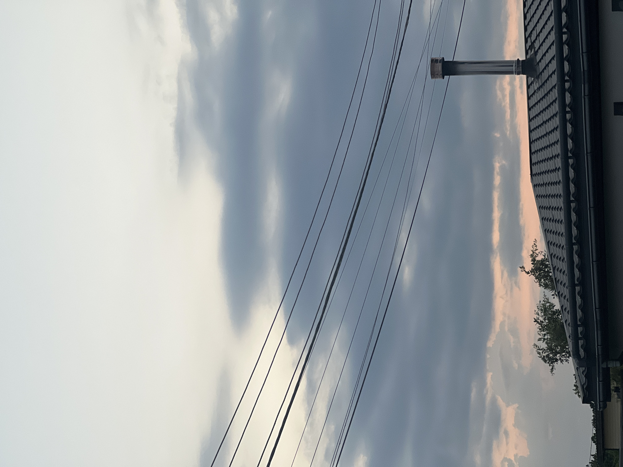 The image is an outdoor, low-angle shot of a cloudy sky with a house roof and power lines in the foreground. The sky is a mix of pale grey and darker, ominous clouds. A silver-colored chimney protrudes vertically from the dark, tiled roof of the house, which takes up the right side of the frame. Several dark power lines run diagonally across the upper portion of the image, disappearing out of frame. A small tree with green leaves is visible behind the roofline. The light suggests it could be late afternoon or early morning, with the clouds creating a muted, possibly overcast or stormy, atmosphere. No people or text are visible in the image.