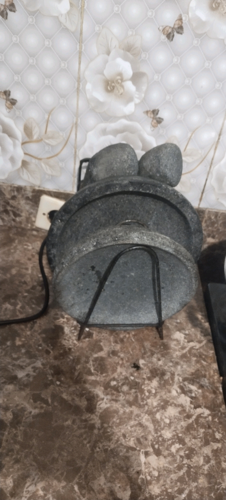 The image shows a stone mortar and pestle set on a marble countertop. The mortar is large and round, with a slightly textured gray surface. Two smaller, rounded stones, presumably pestles, rest on top of the mortar. A black metal stand supports the mortar from underneath. In the background, a tiled wall with a floral pattern and butterflies is visible. To the left of the mortar, a white electrical outlet is mounted on the wall. The overall scene appears to be in a domestic setting, likely a kitchen. There is no indication of the location in Daleman, Indonesia.