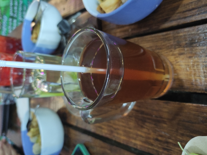 The image captures a close-up, overhead view of several drinks and snacks arranged on a rustic wooden table. The primary subject is a clear glass mug, filled with a dark amber-colored liquid, likely tea, into which a white plastic straw is inserted. The liquid shows a slight translucency.
To the left of the main mug, another clear glass with a lighter liquid, possibly water, is partially visible. Further left, a cup or glass containing a red-colored beverage can be seen. Scattered across the table are several bowls. One prominent blue and white bowl holds a serving of small, light-colored, fried food items, resembling crackers or chips. A similar bowl is partially visible in the upper background.
The setting appears to be a casual dining or relaxation area, likely outdoors or in an establishment with a natural aesthetic, given the sturdy wooden table. The table surface is composed of distinct wooden planks with visible gaps. The overall arrangement suggests a social gathering or a moment of refreshment.
The lighting in the image is soft and even, suggesting either an overcast daytime environment or an indoor setting with diffused light. No specific time of day is discernible, but it does not appear to be nighttime. The focus is sharp on the central drink, with the background elements slightly blurred. There is no visible text identifiable on any of the objects or in the background.