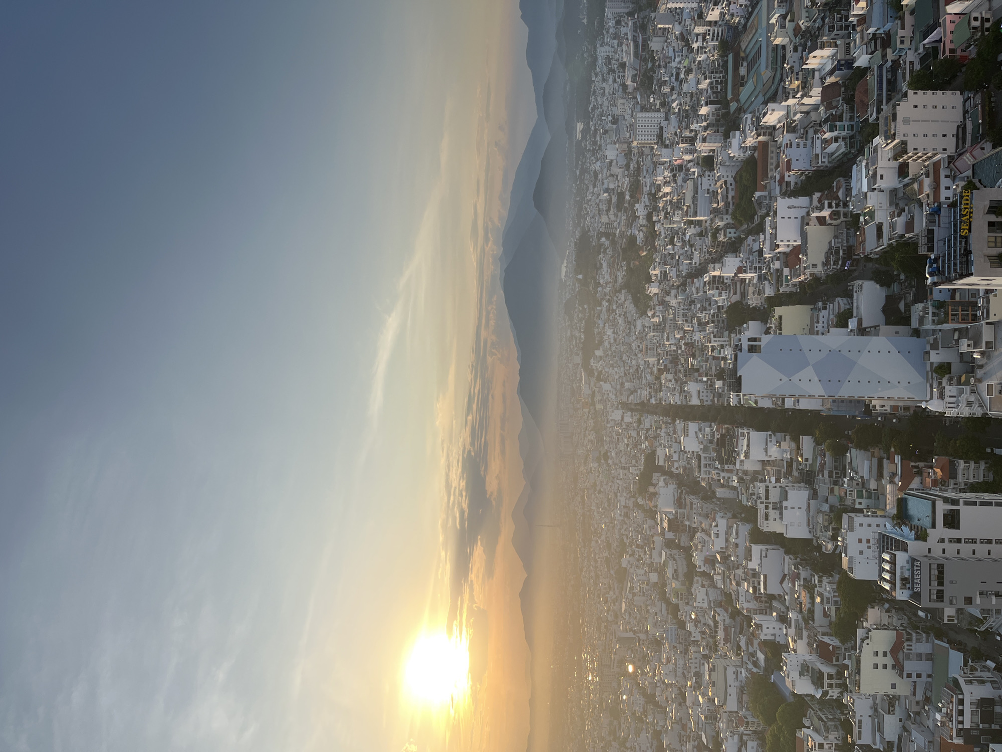 This high-angle, expansive photograph captures a dense urban landscape in Nha Trang Ward, Vietnam, during either sunrise or sunset, indicated by the brilliant sun low on the left horizon. The sun's intense golden light radiates across the sky, transitioning to a pale blue-grey with faint cirrus clouds on the right. In the distant background, a range of hazy, blue-toned mountains forms the horizon line, partially obscured by atmospheric perspective and the ambient light.
The city below is characterized by a high density of low-to-mid-rise buildings, predominantly white or light-colored, spread across a vast area. A prominent, wide thoroughfare extends from the foreground into the mid-ground, bisecting the urban sprawl. Architectural details include numerous flat roofs, some adorned with water tanks or small structures. A distinctive, tall, modern building with a facade featuring a white and light blue geometric, diamond-patterned design stands conspicuously in the lower-middle portion of the image, to the right of the main street. Further towards the bottom, a building with a visible "SEASIDE" sign on its rooftop also features a rooftop swimming pool. Another "SEASIDE" sign is discernible on a building to the left foreground. Green tree canopies are interspersed throughout the cityscape, primarily lining streets and within residential blocks. No individual people or specific actions are discernible due to the elevated perspective.