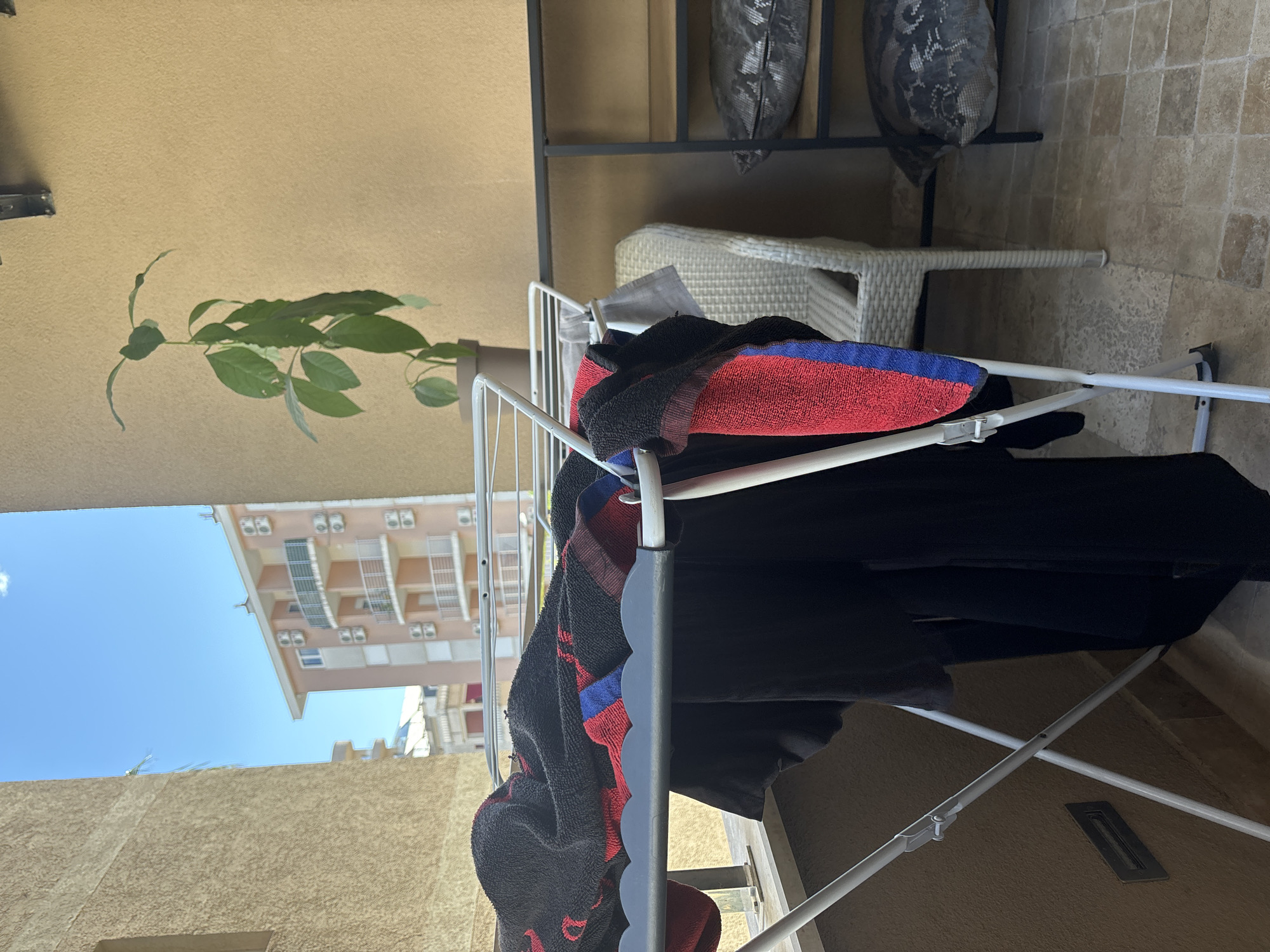The image depicts a clothes drying rack laden with laundry on a balcony in Budva, Montenegro. A dark-colored towel with red and blue accents is prominently draped over the rack. A few other items of dark clothing are also drying. Behind the drying rack, a white wicker chair sits on a tiled balcony floor. A black metal shelf unit is visible in the background, with what appear to be gray patterned pillows on the shelf. The photograph is taken at an angle, partially obscuring the view of the city outside. The sky is blue, and a multi-story peach-colored building is seen in the distance. The balcony itself has a tan-colored wall. A green leafy plant stands in a pot next to the drying rack.