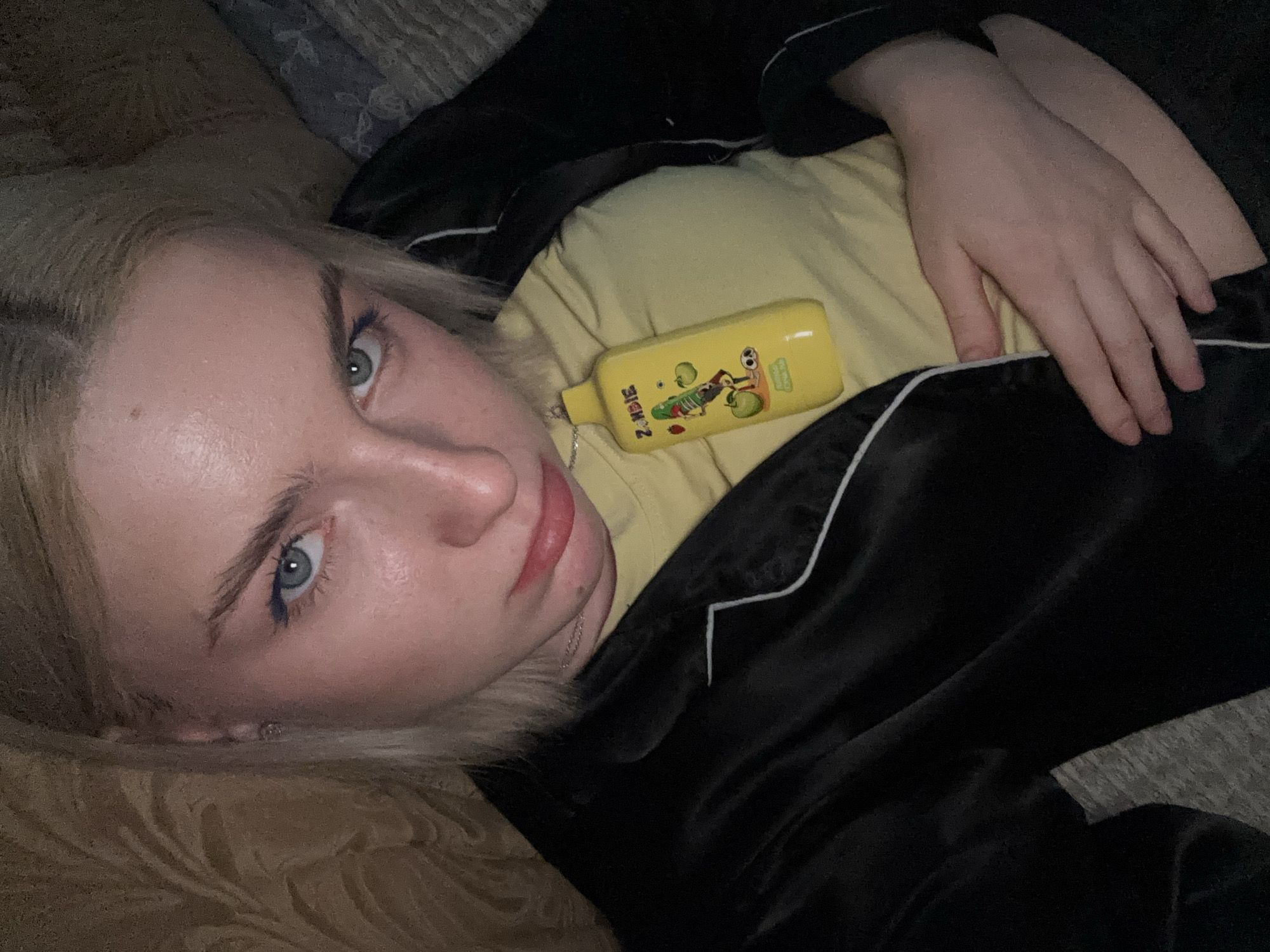 A young woman with blonde hair and blue eyes is lying on her side, looking at the camera. She is wearing a yellow shirt and black pajamas with white piping. A small, yellow, rectangular object with cartoon characters and the word "ZOMBIE" is resting on her chest. Her left hand is placed over the object, and her right arm is bent, with her hand resting on her shoulder. The background appears to be a bed or sofa with a textured surface. The lighting is dim, suggesting it might be nighttime or indoors. The woman's expression is neutral, with a slight pout. The overall mood is relaxed and intimate. The word "ZOMBIE" is clearly visible on the object.