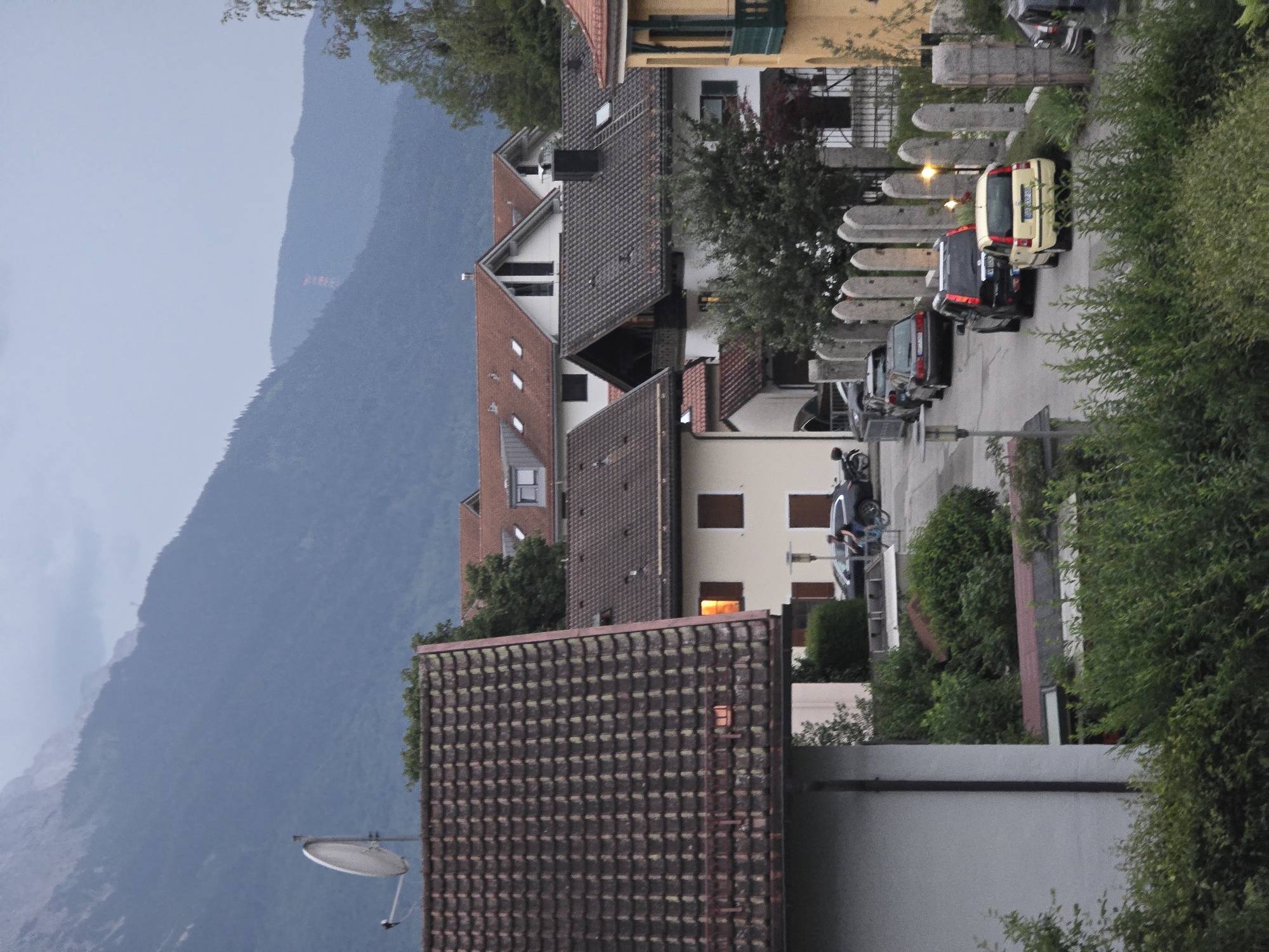 The image depicts a street scene in Garmisch-Partenkirchen, Germany, with a backdrop of a large, tree-covered mountain under a cloudy sky. The foreground features dense green foliage. A road curves through the scene, lined with several houses with tiled roofs and windows.
Several cars are parked along the street. A yellow Fiat 500 is prominently positioned in the middle of the road, facing away from the viewer. To its left, a black SUV is parked, and further back, a dark sedan is visible. A person is riding a bicycle on the right side of the road, moving away from the camera.
Other objects in the scene include a satellite dish mounted on a rooftop, stone pillars lining a property, and a street lamp. The architecture of the houses suggests a traditional Bavarian style. The overall impression is that of a quiet residential area on an overcast day.