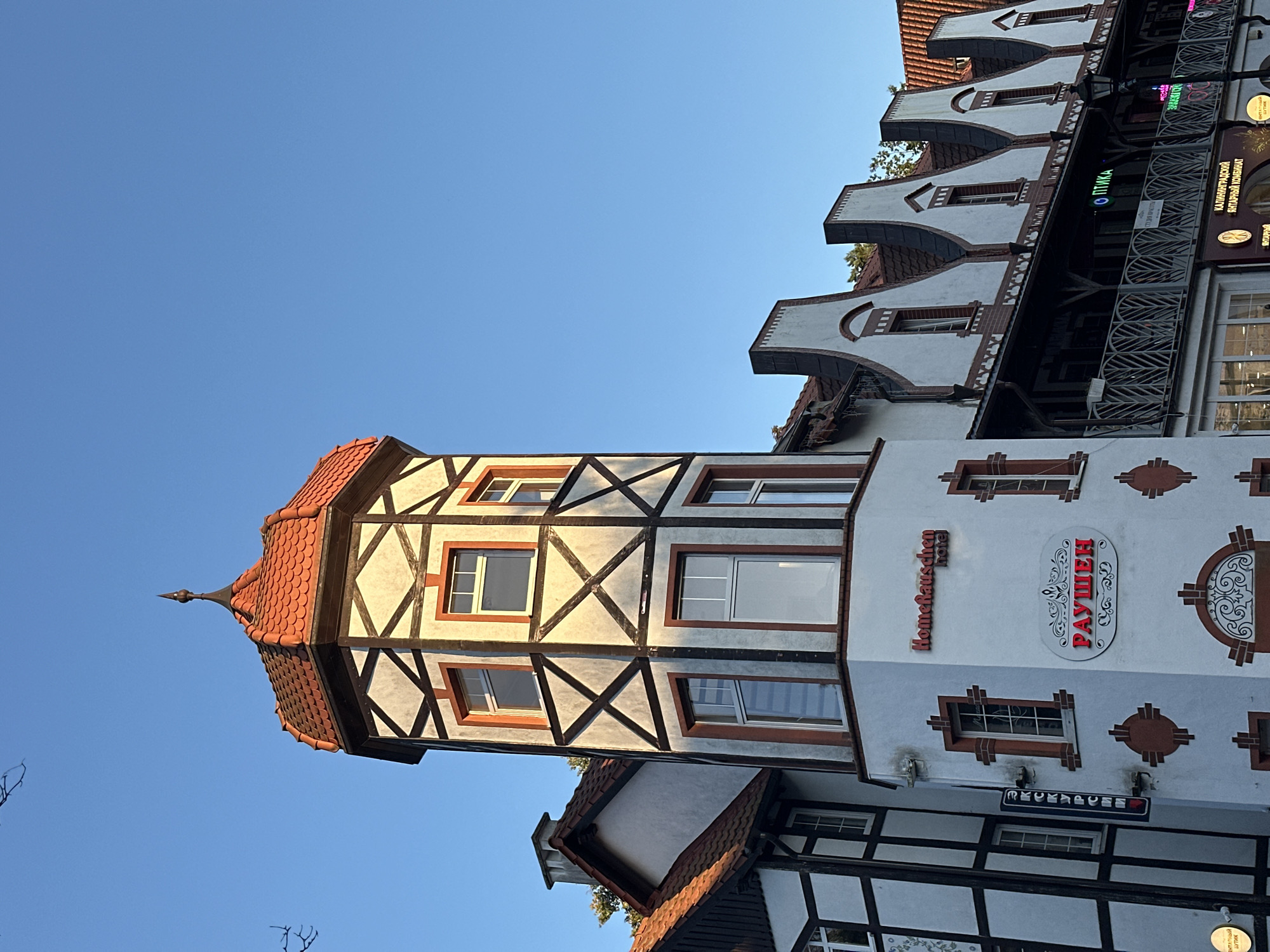 The image shows a multi-story building with a distinctive red-tiled, octagonal turret capped with a dark spire. The turret has exposed timber framing in a dark crisscross pattern against a light-colored facade, with orange trim around the windows. Adjacent to the turret, a row of buildings features a similar architectural style with pointed gables, dark timber accents, and red tile roofing. Balconies with decorative metal railings run along the upper floors. Signs on the building include "HomeRauschen hotel" and "РАУШЕН" in Cyrillic script, indicating this is likely the "Rauschen" hotel in Svetlogorsk, Russia, a resort town known for its German-influenced architecture. The sky is clear and blue, and the lighting suggests it is late afternoon or early morning, casting a warm glow on the building. There are no people visible in the image.