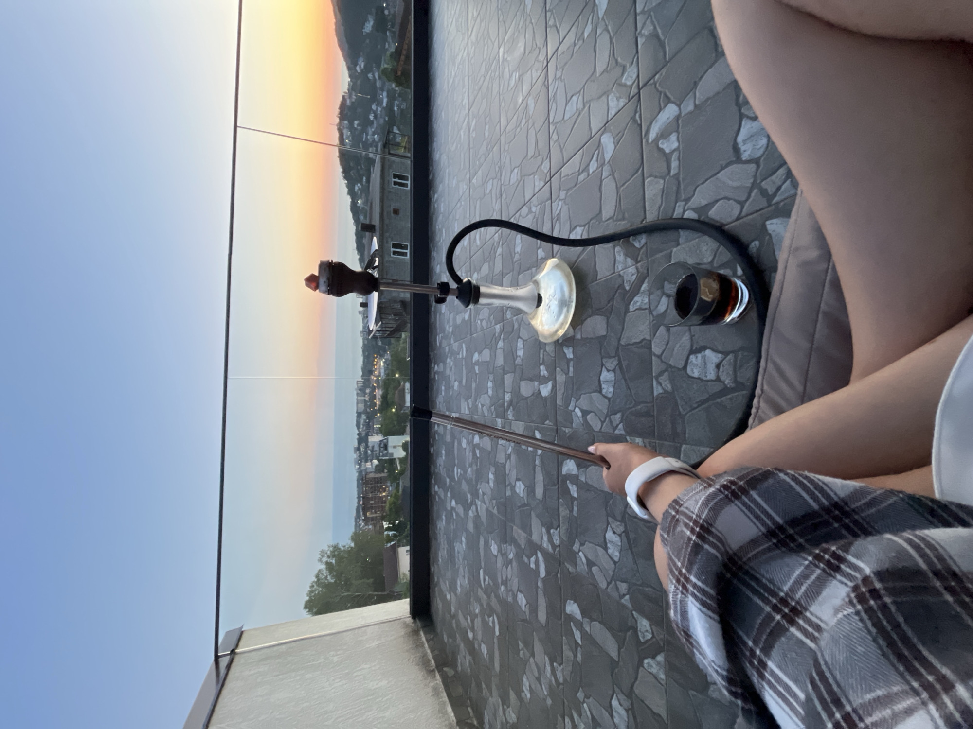 A high-angle, first-person perspective photograph captures a scene on an elevated terrace in Baranovka, Russia, during twilight. The lower body and left arm of an individual, seated on a light grey cushion, are visible. The individual wears a grey and white plaid garment and a white wristband on the left wrist. The left hand grasps the mouthpiece of a black hose attached to a clear-based, dark-stemmed hookah, which rests on the patterned grey and white tiled floor. Glowing coals are present in the hookah's bowl. Adjacent to the hookah, a clear glass contains a dark liquid with a yellow citrus wedge. A glass and metal railing demarcates the terrace edge, providing an unobstructed view of a coastal city and mountainous landscape under a gradient sky transitioning from blue to orange at the horizon. The Black Sea is discernible in the distance. Urban structures and developing illumination are visible across the city and ascending into the vegetated hills.