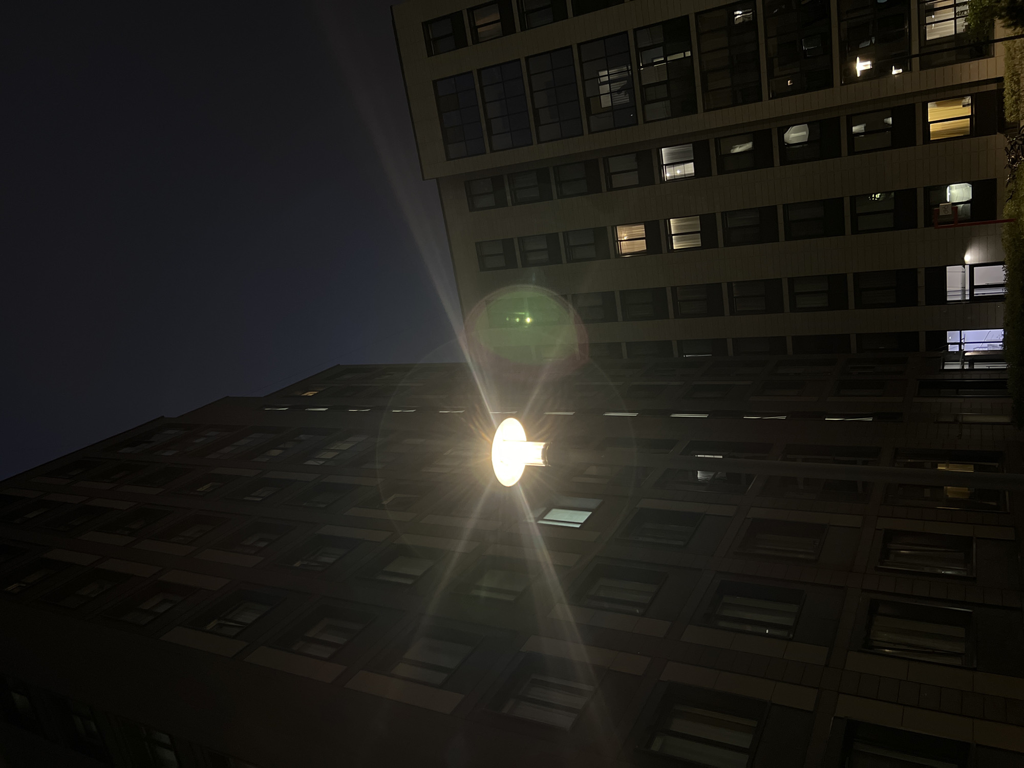The image shows a night view of a courtyard in a residential complex in Saint Petersburg, Russia. Two large apartment buildings are visible, one on the left and another on the right, both with multiple floors and numerous windows. A street lamp is prominently featured in the center of the image, casting a bright light and creating lens flare. Some windows in the buildings are illuminated, suggesting that people are inside. The sky is dark, indicating it is nighttime. The architecture of the buildings appears modern.