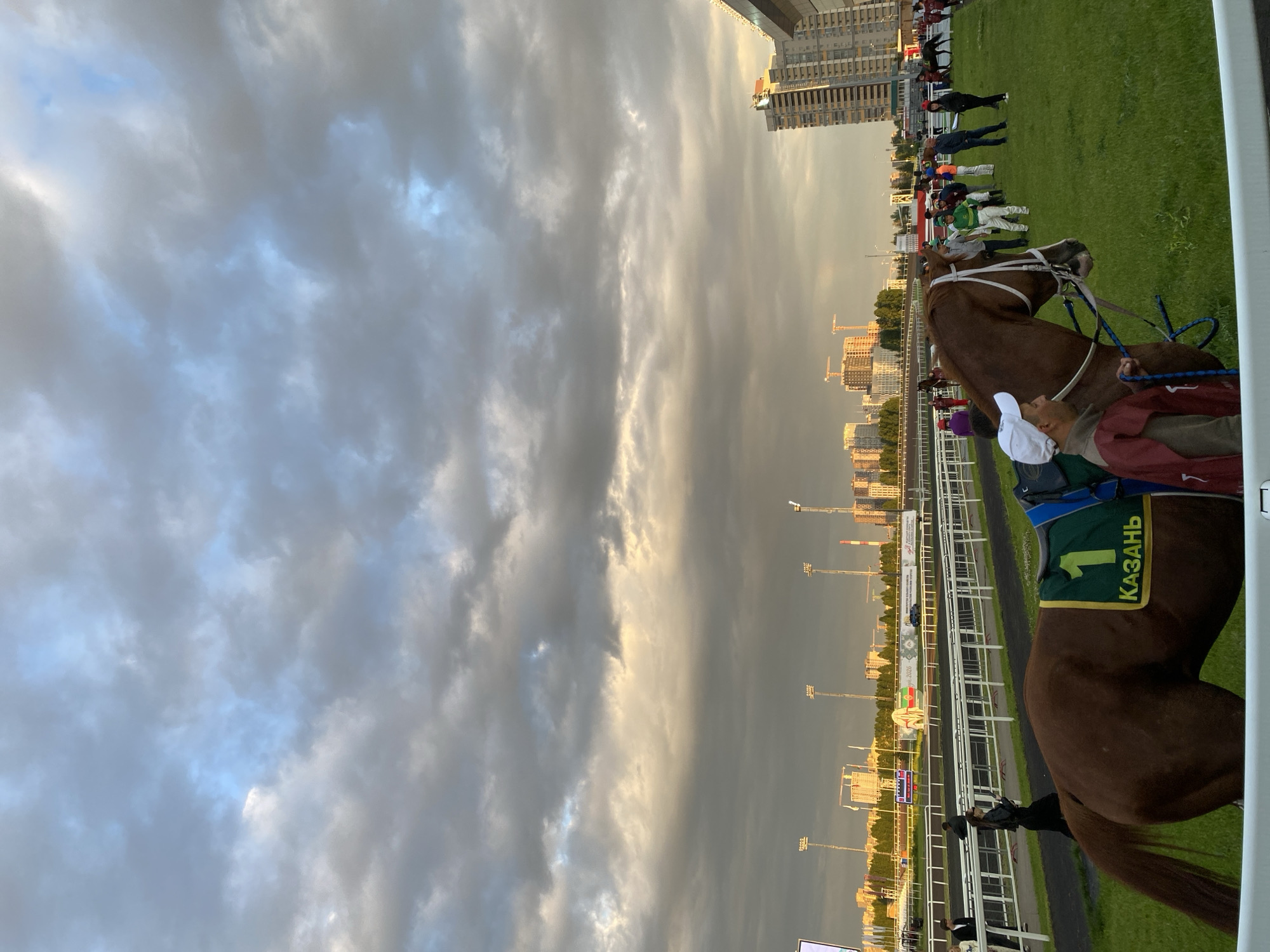 The image depicts a horse race event at the Kazan Hippodrome, located in Kazan, Russia. The foreground features a brown horse with a green saddle blanket bearing the number "1" and the inscription "КАЗАНЬ". A person wearing a white cap and a maroon vest is holding the horse's reins. Several other individuals, including jockeys in racing attire and spectators, are visible on the grassy field and near the track. In the background, the Kazan skyline is visible, with tall buildings and construction cranes under a dramatic, cloudy sky. The race track, lined with white fences, stretches into the distance. The overall scene captures a moment before or after a race, with a sense of anticipation and the urban landscape providing a backdrop.
