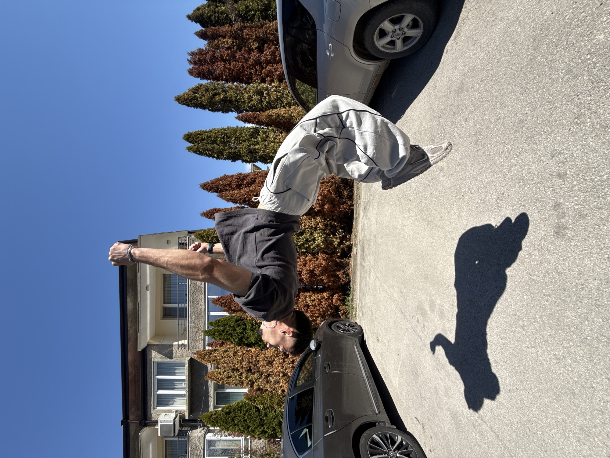 A man is performing a backflip on a street in Shumen, Bulgaria. He is wearing a dark gray t-shirt, light gray sweatpants, and gray sneakers. His arms are raised and extended straight upward above him, toward the upper-left corner of the image. He is located between a parked gray sedan and a silver hatchback. Adjacent to these vehicles is a row of conifer trees, some of which are brown, and a tan-colored building with white-framed windows. The sky is clear and blue, and there is a prominent shadow on the pavement cast by the backflipping man.