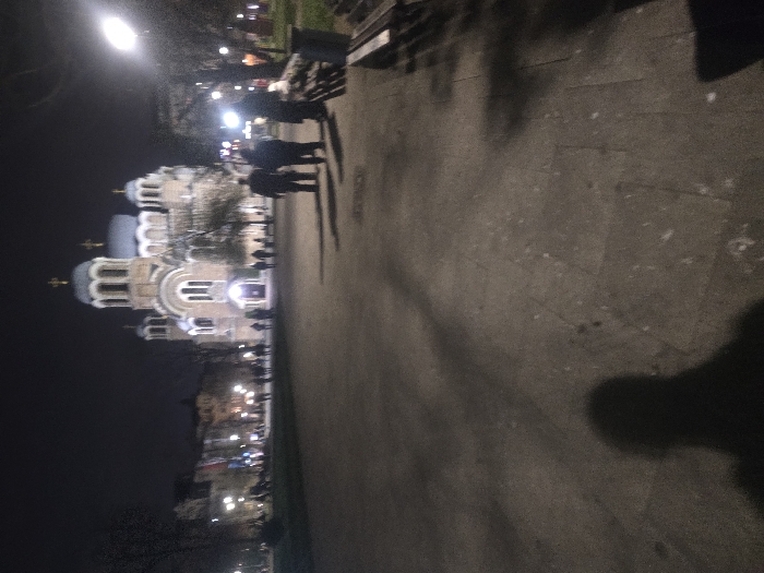 The image displays the Sveti Sedmochislenitsi Church at night, situated in an urban park in Sofia, Bulgaria. The church, a prominent Orthodox structure featuring multiple domes topped with crosses, is illuminated in the background. A paved walkway extends from the foreground into the midground. Several individuals are present: three persons are depicted from behind in the midground, casting long shadows to the left, and additional figures are discernible closer to the church's entrance. On the far right, a bright streetlamp emits light, and a dark park bench with a waste bin is partially visible. The immediate foreground consists of paved ground, displaying visible seams and the distinct, dark shadow of the photographer. The overall environment is a park setting with visible bare trees, under a dark night sky with artificial lighting.