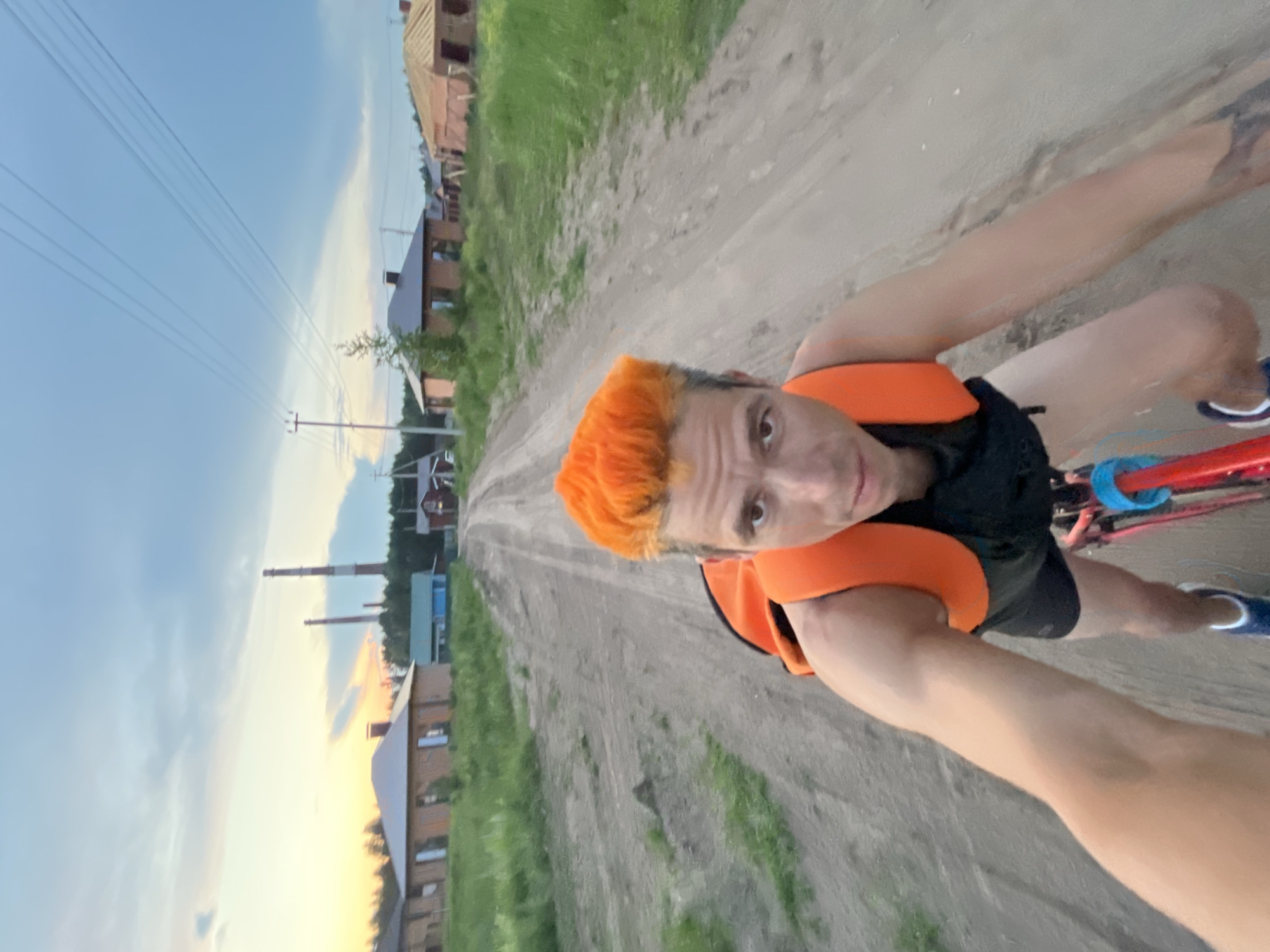 A person with bright orange hair rides a bicycle down a dirt road in a suburban area. The person is wearing an orange backpack. The road is surrounded by green grass and several houses. In the background, there are two smokestacks and power lines. The sky is cloudy with the sun setting on the horizon, casting a yellow and orange glow. The location is identified as Chaikovsky, Russia.