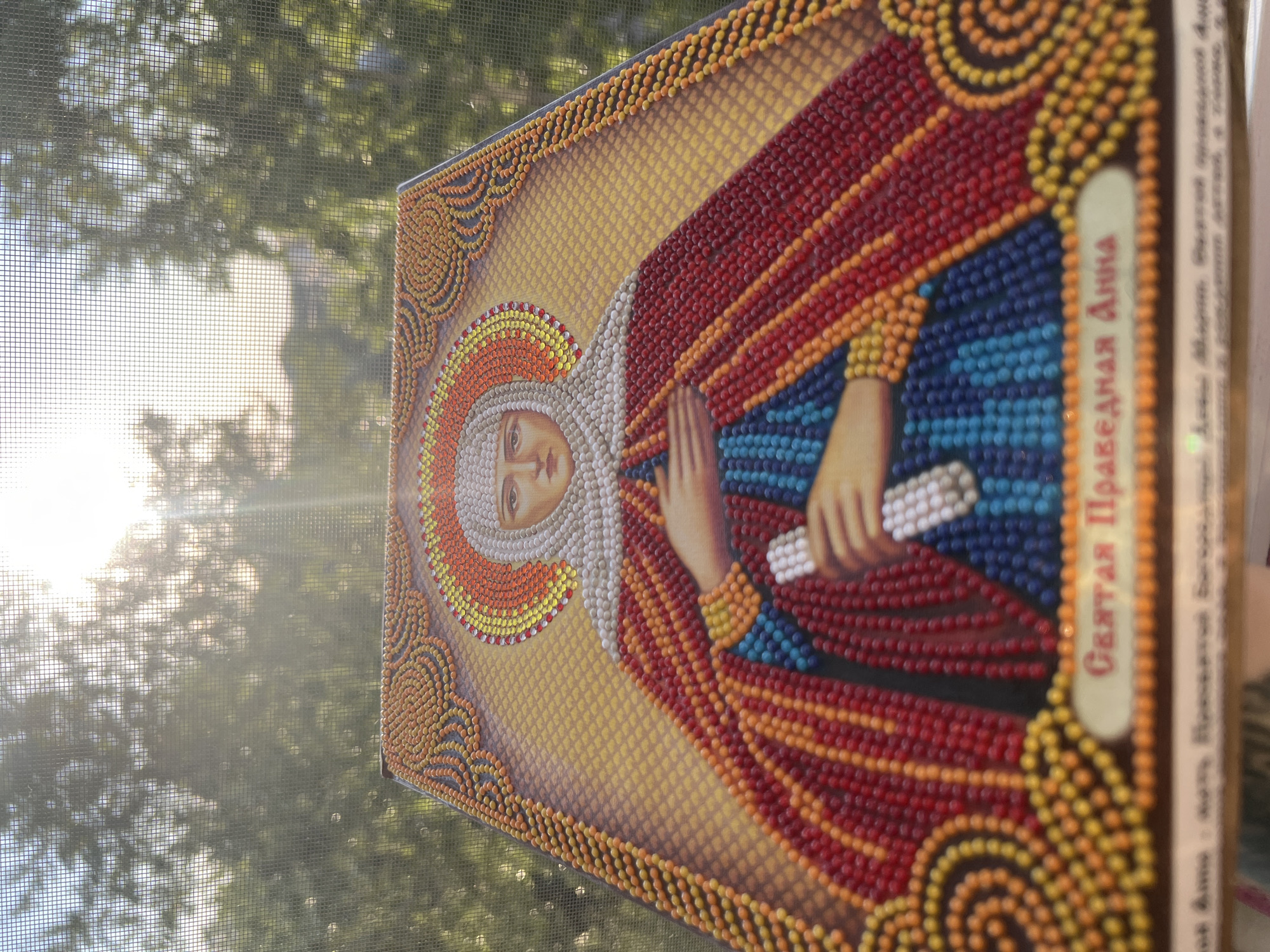 The image shows a close-up, slightly angled view of a diamond painting of an Orthodox icon. The icon depicts a female saint with a halo, wearing a blue and red garment and a white veil. She is holding a white object in her hand. The painting is made of small, colorful beads arranged in a mosaic pattern. The background of the painting is a gradient of yellow and orange, with decorative borders. Below the saint's image, there is text in Cyrillic script.
The diamond painting is placed in front of a window with a screen. Sunlight filters through the screen, creating a hazy effect and illuminating the scene. Through the screen, trees and the sky are visible, suggesting an outdoor or semi-outdoor setting. The overall lighting is bright and diffused. The lower edge of the diamond painting is visible, along with a portion of what appears to be its packaging or frame, which also has some Cyrillic text. The image appears to be taken indoors, looking out through a window. There is no indication of the city of Omsk, Russia within the image itself, nor any environmental cues that would allow for its identification.