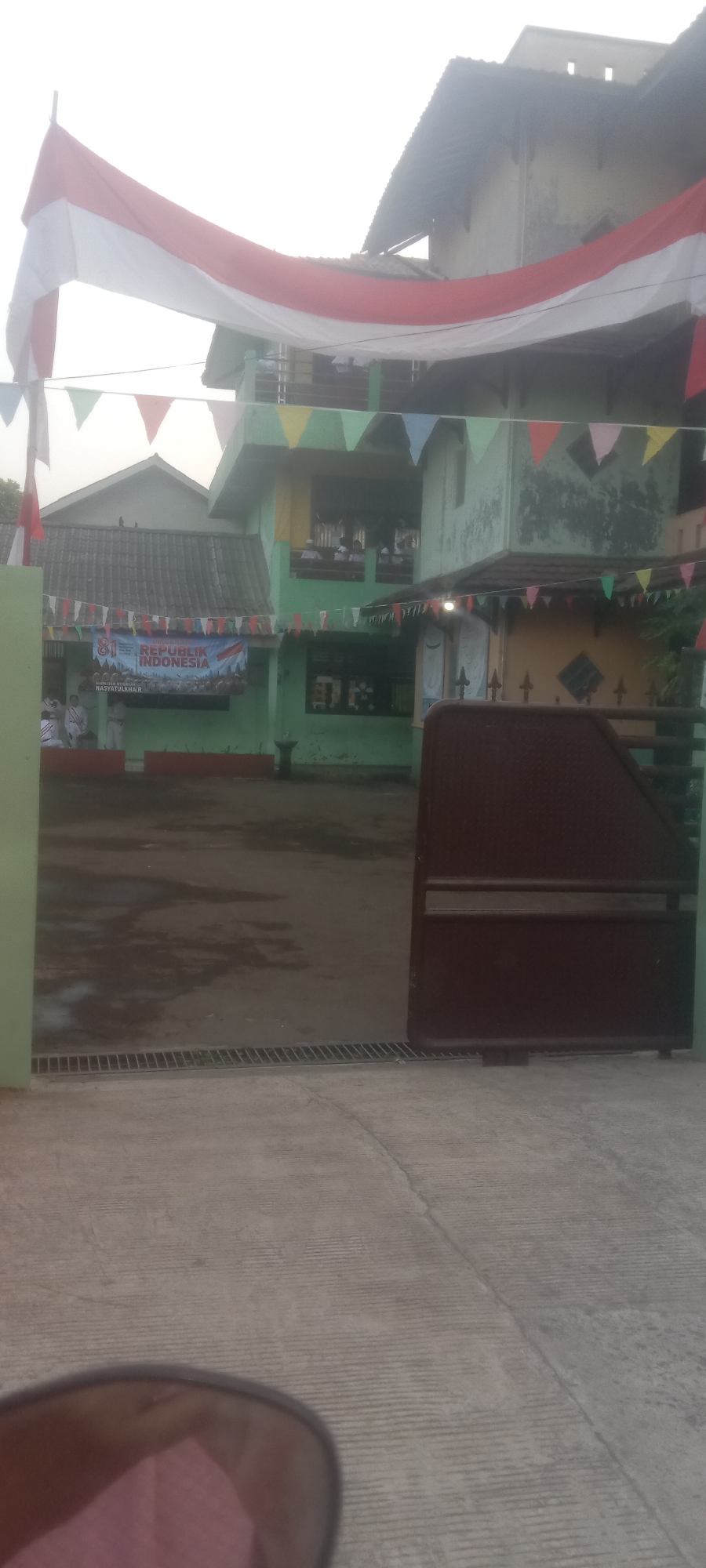 This image captures the exterior of a light green building, likely a school or community center, in Depok, Indonesia. The building has multiple stories and is decorated for a national celebration, possibly Independence Day, with a large Indonesian flag draped across the front and colorful triangular flags strung along the balconies and rooflines. A banner on the lower level reads "81 REPUBLIK INDONESIA" and "HASYATULHAIR". Several people, appearing to be students in uniform, are visible on the balconies and in front of the building. The ground in front of the building is paved and wet, suggesting recent rain. A dark brown metal gate is partially open on the right side of the frame, and a portion of a concrete walkway with a metal grate is visible in the foreground. The overall atmosphere appears festive and communal.