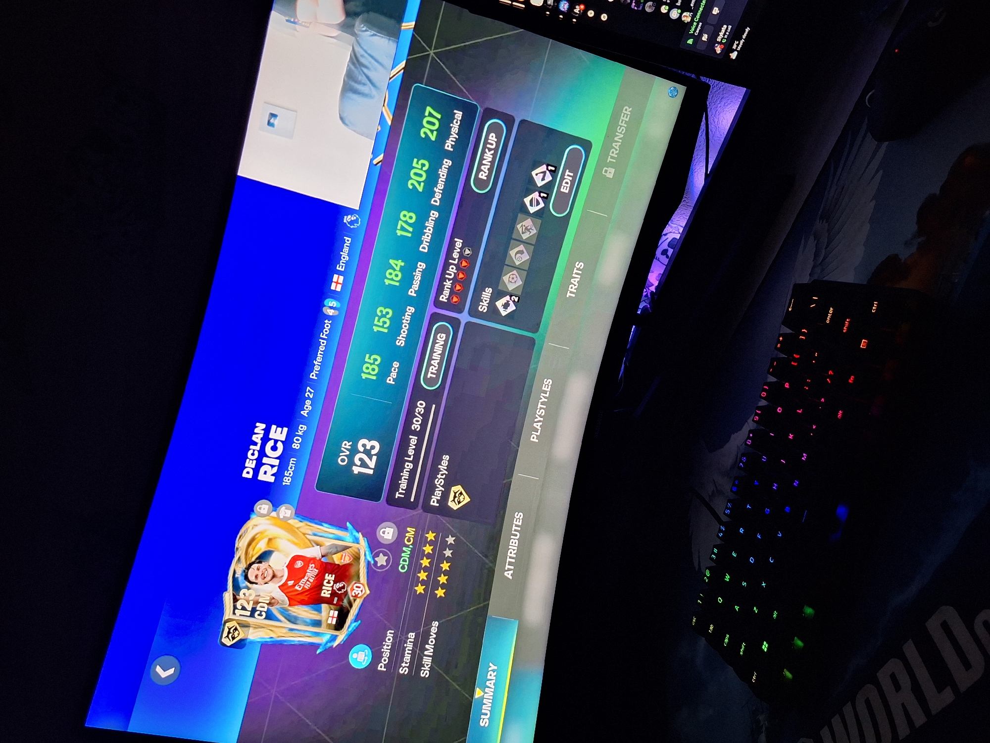The image shows a close-up, eye-level, indoor shot of a curved computer monitor displaying a video game interface, specifically a player card for "DECLAN RICE." The player card shows his stats, including an overall rating of 123, and attributes such as Pace, Shooting, Passing, Dribbling, Defending, and Physical. The player's image features him in an Arsenal football kit. Below the player card, there are tabs for "SUMMARY," "ATTRIBUTES," and "PLAYSTYLES." The bottom right of the monitor displays a portion of a chat interface with user avatars and names.
In the foreground, a black mechanical keyboard with colorful RGB backlighting is visible, resting on a desk mat that appears to have a mountain or cloud design. A computer mouse is partially visible to the right of the keyboard. The environment appears to be a dimly lit room, with the monitor's blue and purple lighting casting reflections on the surroundings. There is no information in the image to suggest a location in Varna, Bulgaria.